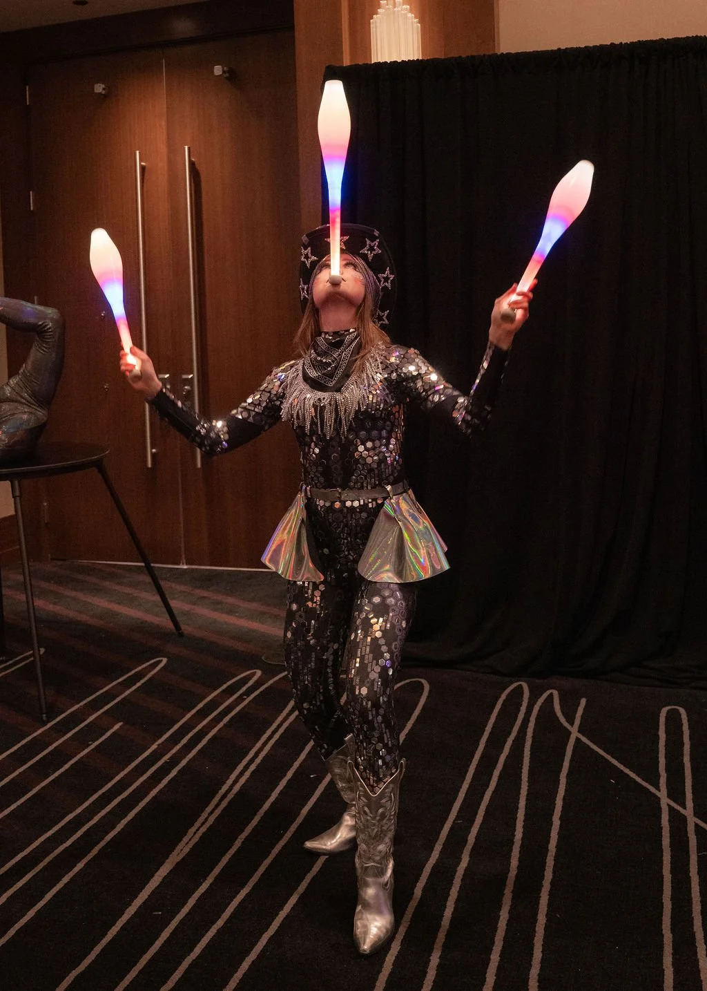 A performer in a sequined costume with holographic ruffled shorts, silver boots, and a starry hat juggles three lit juggling clubs in a dark curtain background.