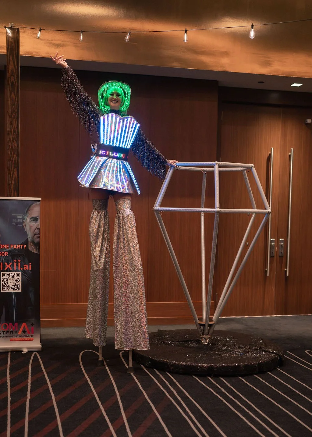A performer on stilts dressed in a shiny, colorful costume with neon lights, green wig, and glittery pants, standing next to a geometric metal sculpture inside a room with wood-paneled walls and string lights overhead.