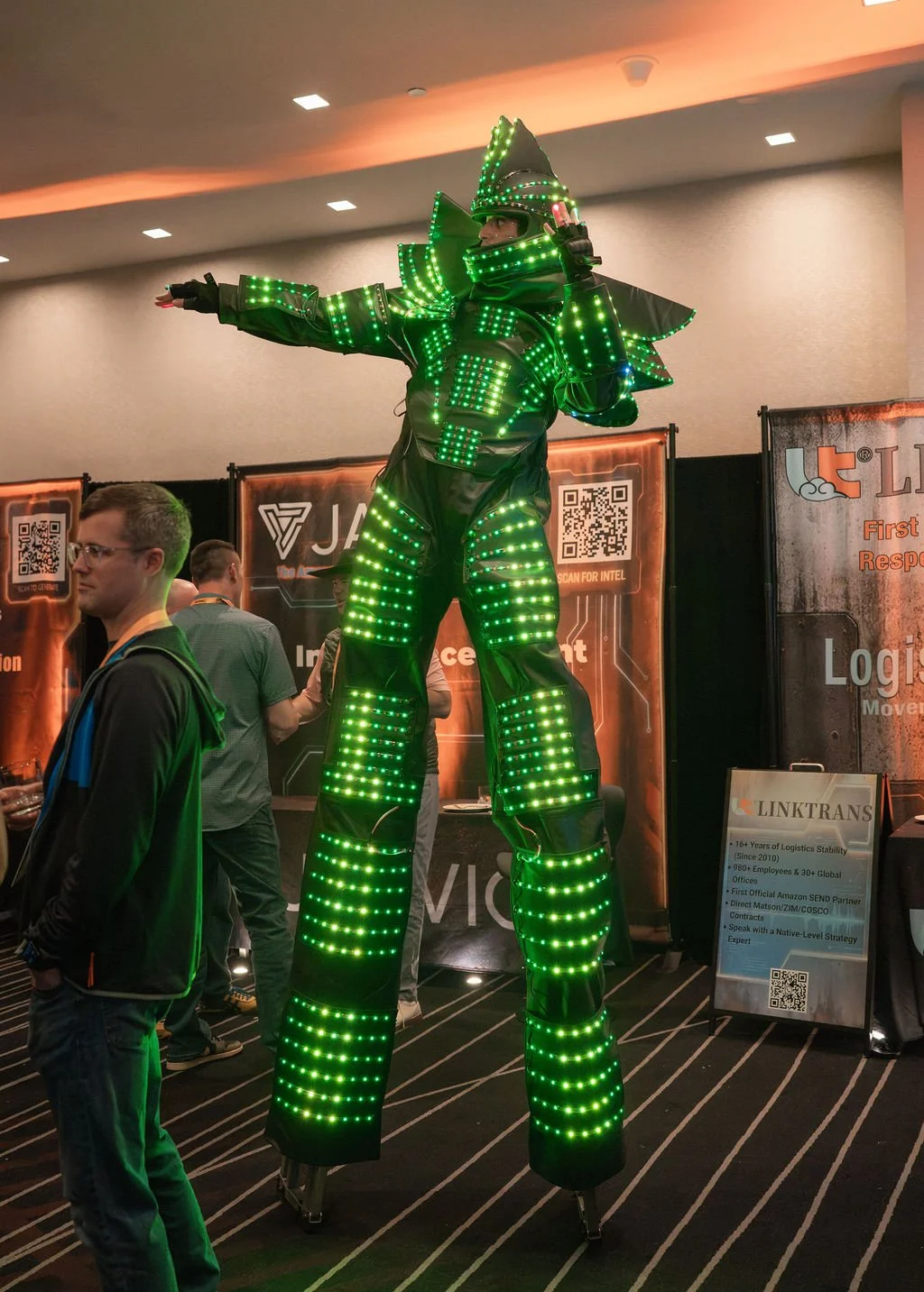 A costumed performer on stilts dressed in a futuristic, LED-lit outfit with a witch hat, illuminated in green lights, posing at a convention or event.