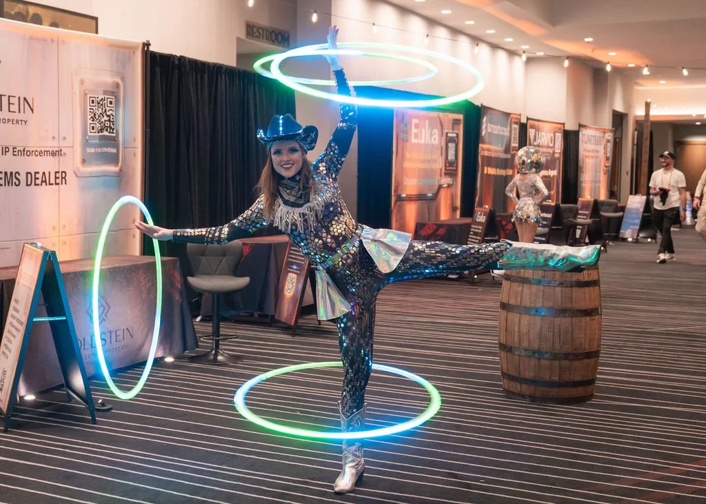 A woman dressed as a circus performer or entertainer performing with glowing hula hoops and metallic costume in an indoor convention center or event space.