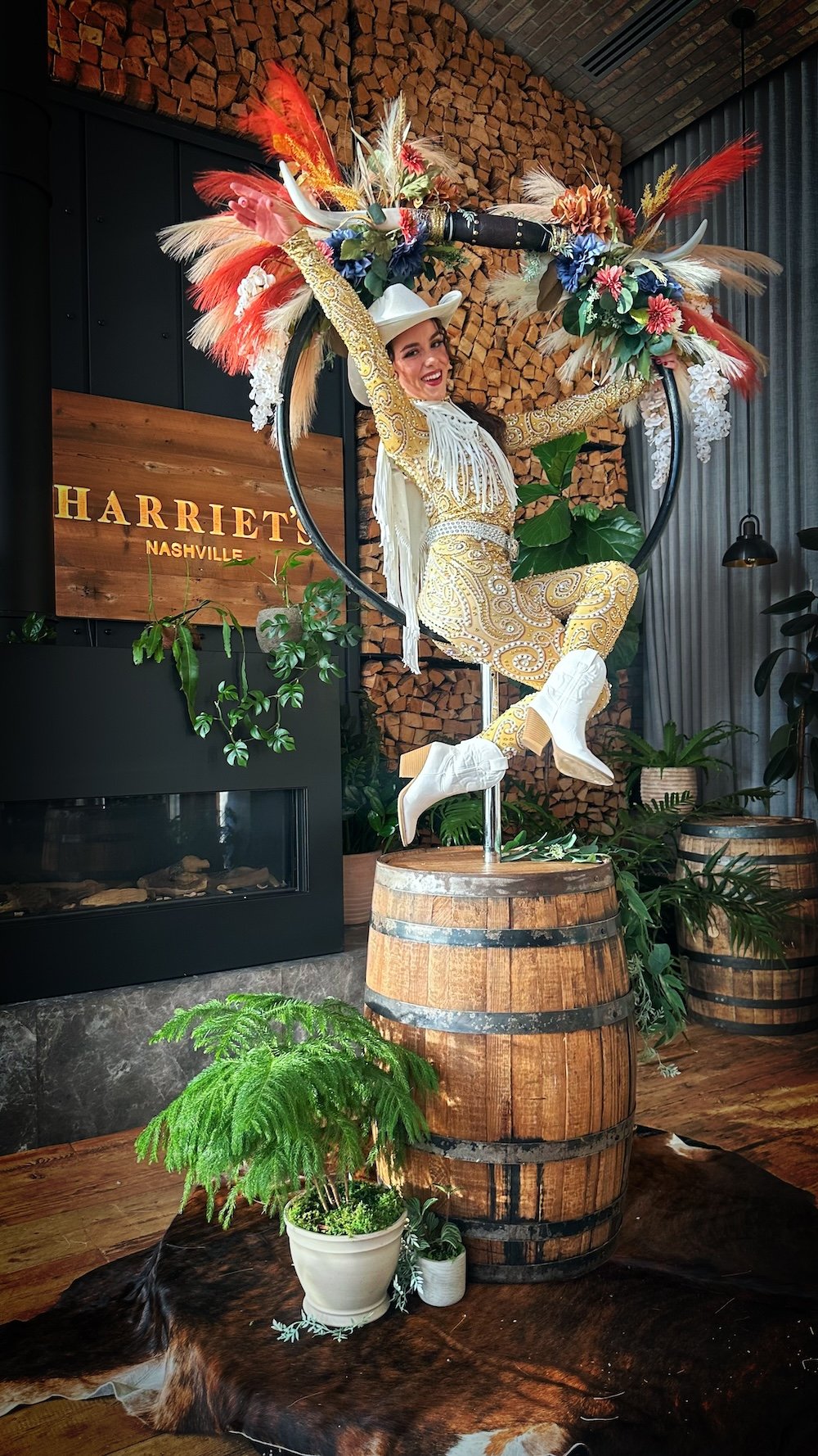 Aerialist in a gold sequined fringe Western costume, white cowboy hat and boots seated in a floral-crowned aerial hoop on a whiskey barrel at Harriet's Rooftop in Nashville