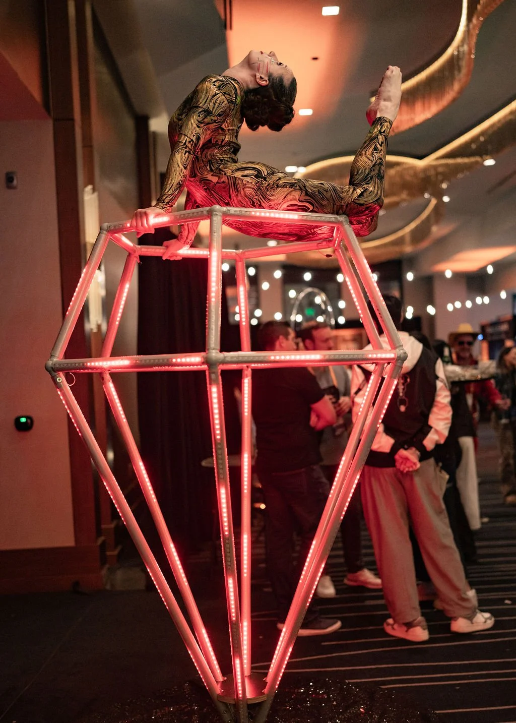 A performer with body paint and long hair, posing atop a large illuminated diamond-shaped sculpture with red LED lights, in an indoor event space with people in the background.