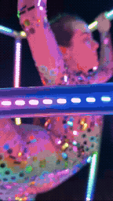 Close-up of a person with short hair and colorful glitter makeup, wearing a multicolored, embellished top, with neon lights in the background.
