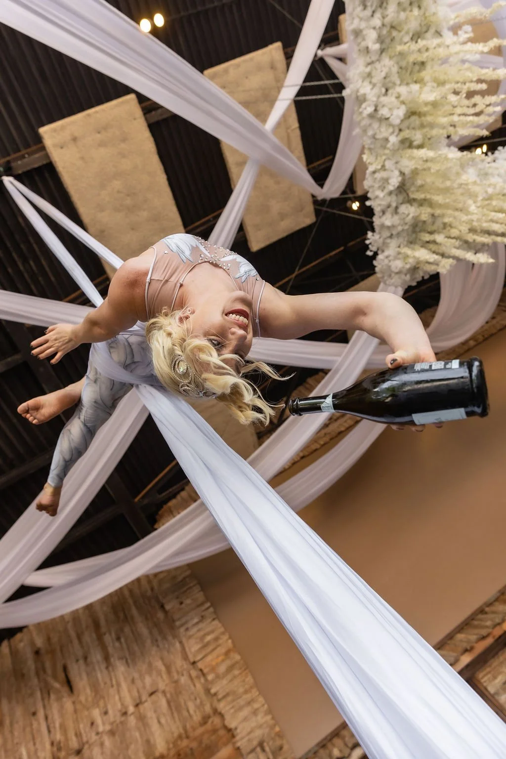 A woman hanging upside down holding a champagne bottle at a celebration or party, with decorations including white fabric drapes and floral arrangements overhead.
