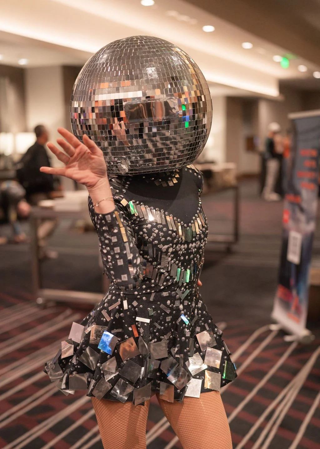 Person wearing a disco ball helmet and a black dress decorated with shiny silver and iridescent square and rectangular mirrors, waving at the camera in an indoor event space.