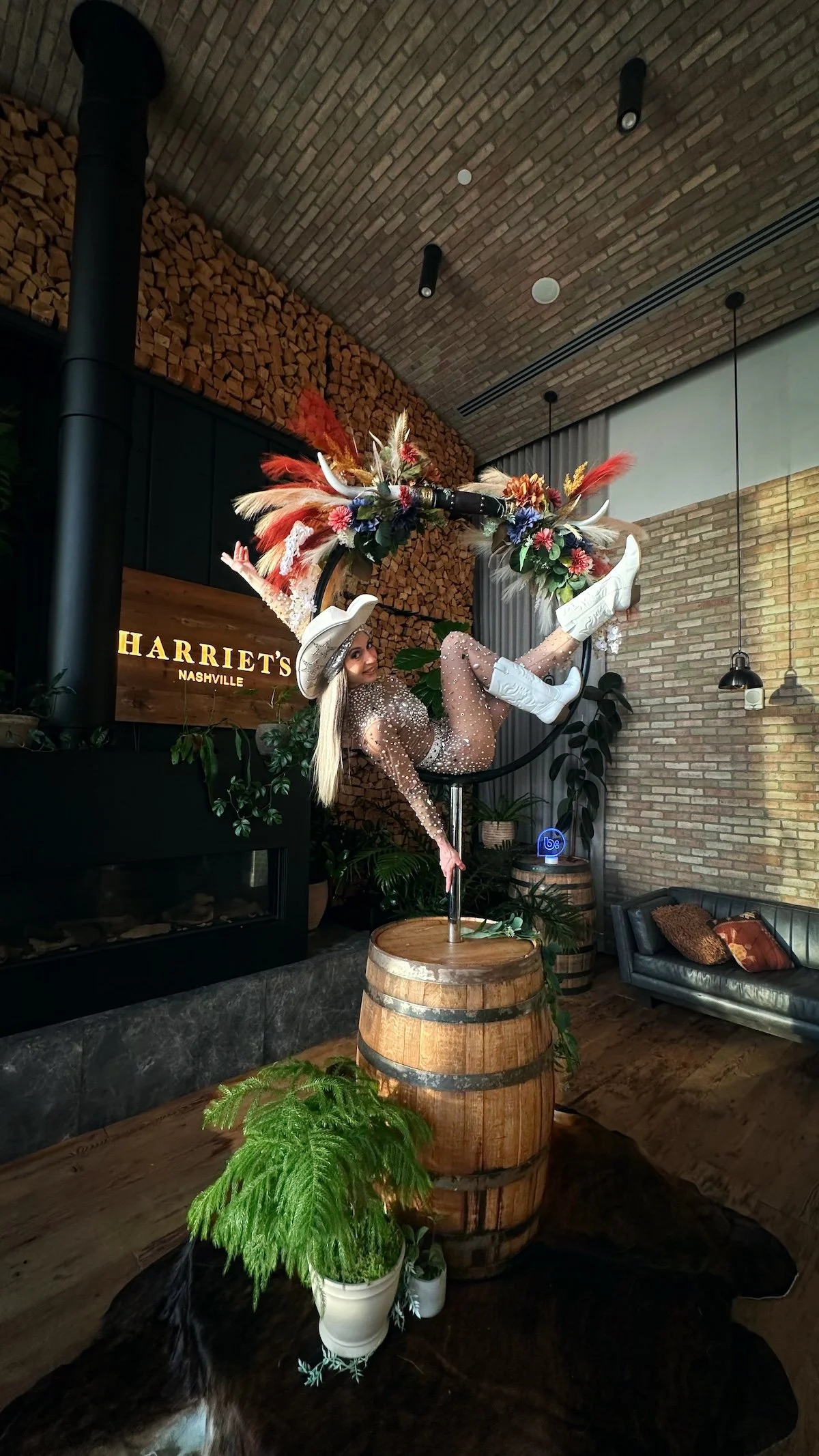 Aerialist in a rhinestone bodysuit, white cowboy hat and boots posing in an aerial hoop crowned with pampas, florals and longhorns, mounted on a whiskey barrel at Harriet's Rooftop in Nashville