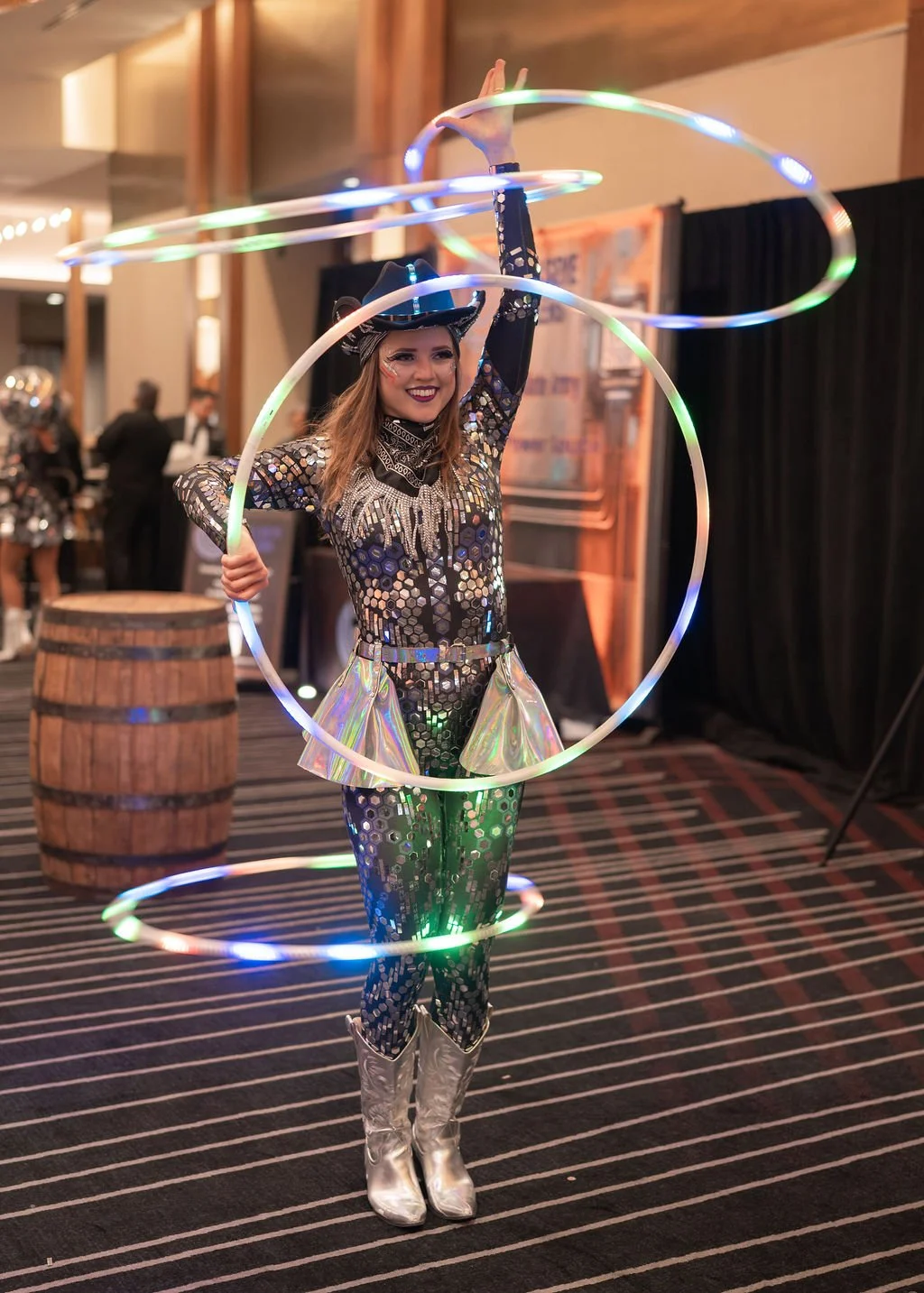 A woman performing a hula hoop act, dressed in a shiny, patterned costume with metallic silver boots, at an indoor event.