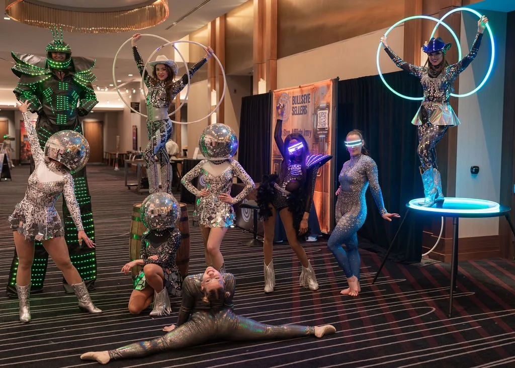 Group of performers dressed in futuristic, sparkling costumes with some wearing LED outfits and others with disco balls on their heads, posing indoors with LED hula hoops and a high-tech robot-like figure.