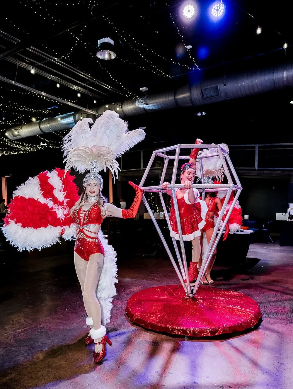 Performers dressed in red and white costumes with feathered headdresses and large props, performing at a festive event with string lights and stage lights overhead.