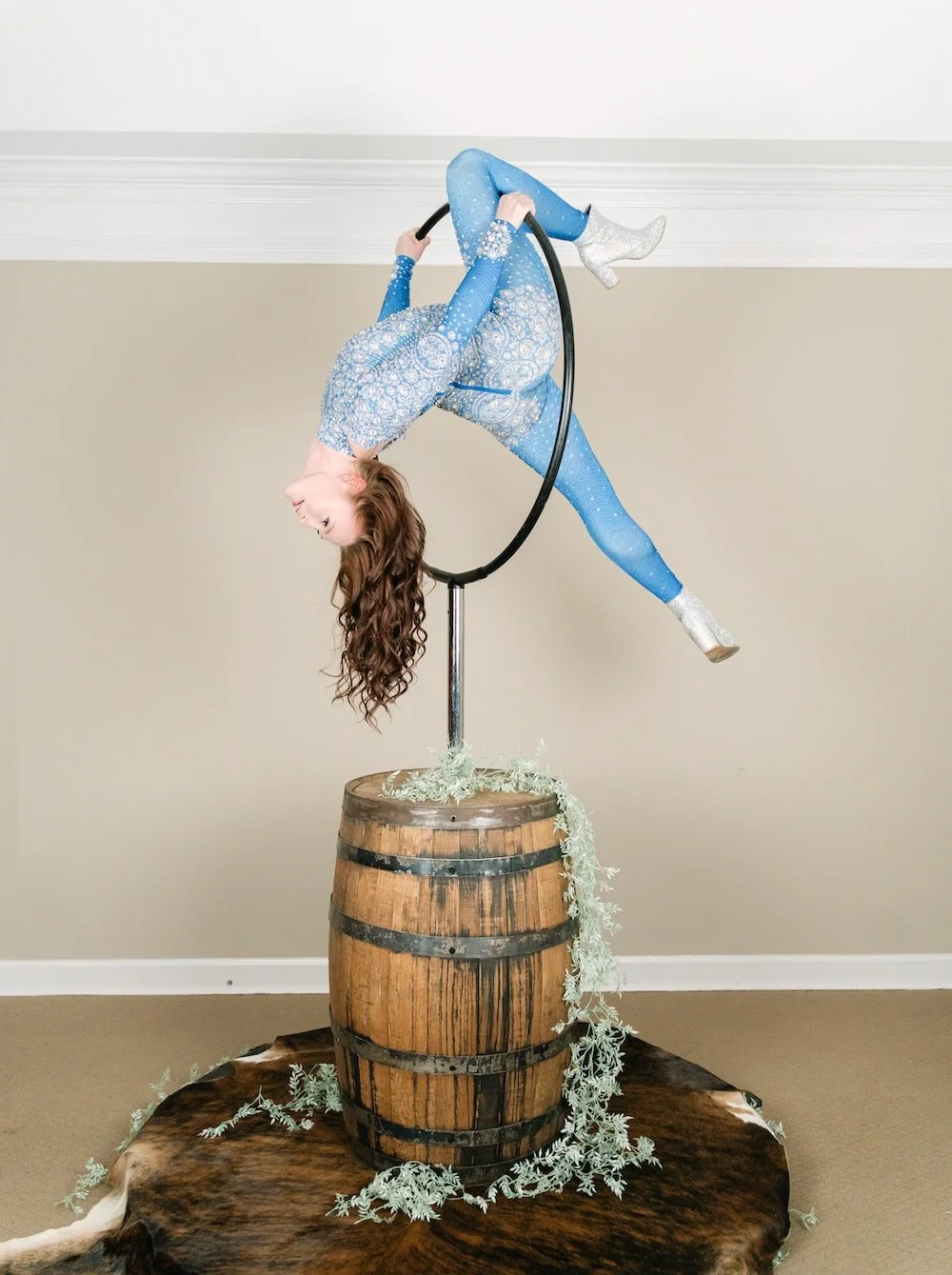 Aerialist in a blue patterned bodysuit hanging upside down from an aerial hoop mounted on a whiskey barrel, styled with trailing greenery on a cowhide base