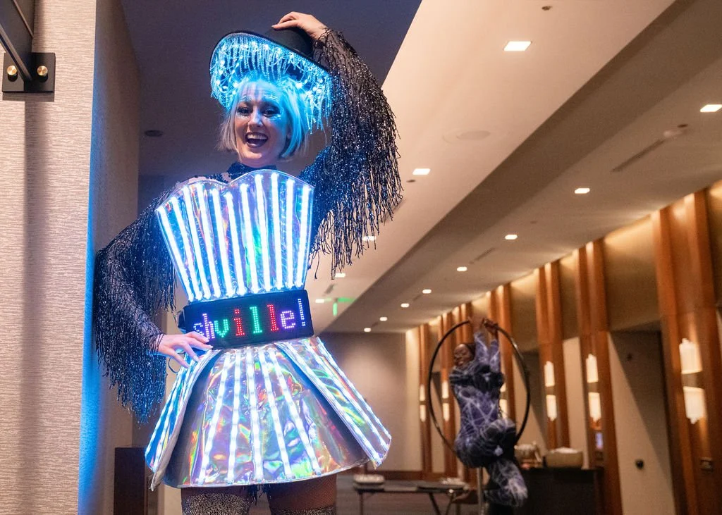 A woman in a futuristic, illuminated costume with the word 'souville' on her waist, smiling and posing with her hand on her hat in a modern indoor setting.