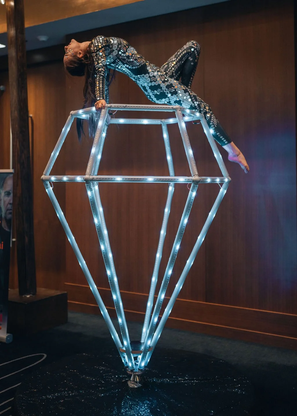 A woman in a shiny, patterned bodysuit performing a balancing act on a geometric illuminated structure in an indoor setting.