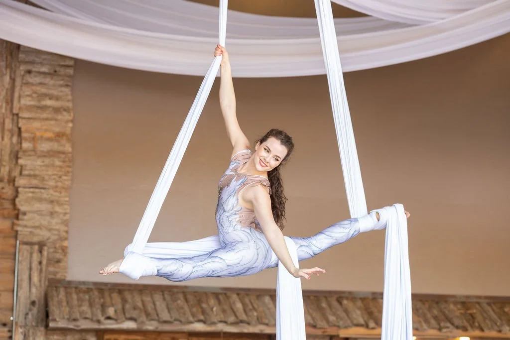 A woman performing aerial silk acrobatics indoors, wearing a patterned outfit and smiling.