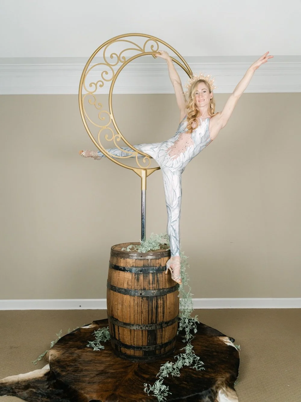 Aerialist in a white lace costume balancing in an arabesque on a gold crescent-moon aerial rig mounted on a whiskey barrel over a cowhide rug