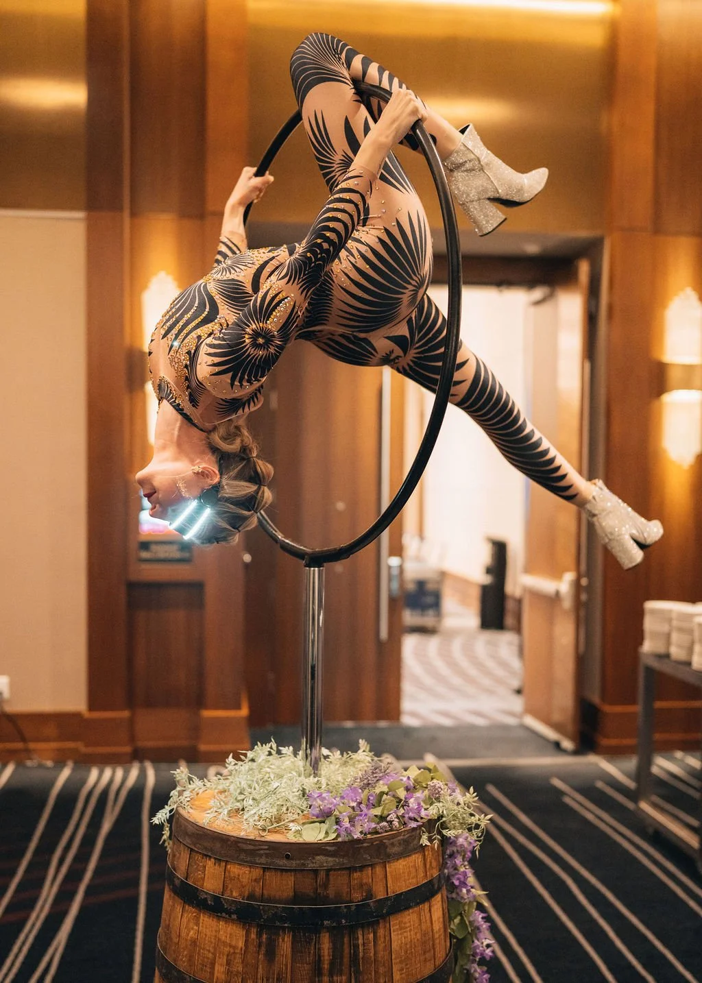 A woman performing an acrobatic act inside a metal ring on a wooden barrel with flowers, wearing a black and gold patterned bodysuit and glittery silver high heels, in a room with wood-paneled walls.