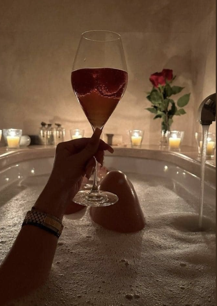 Person holding a glass of red wine over a bubble bath with a heart-shaped bath bomb, candles, and a single red rose in a bathroom with soft lighting.