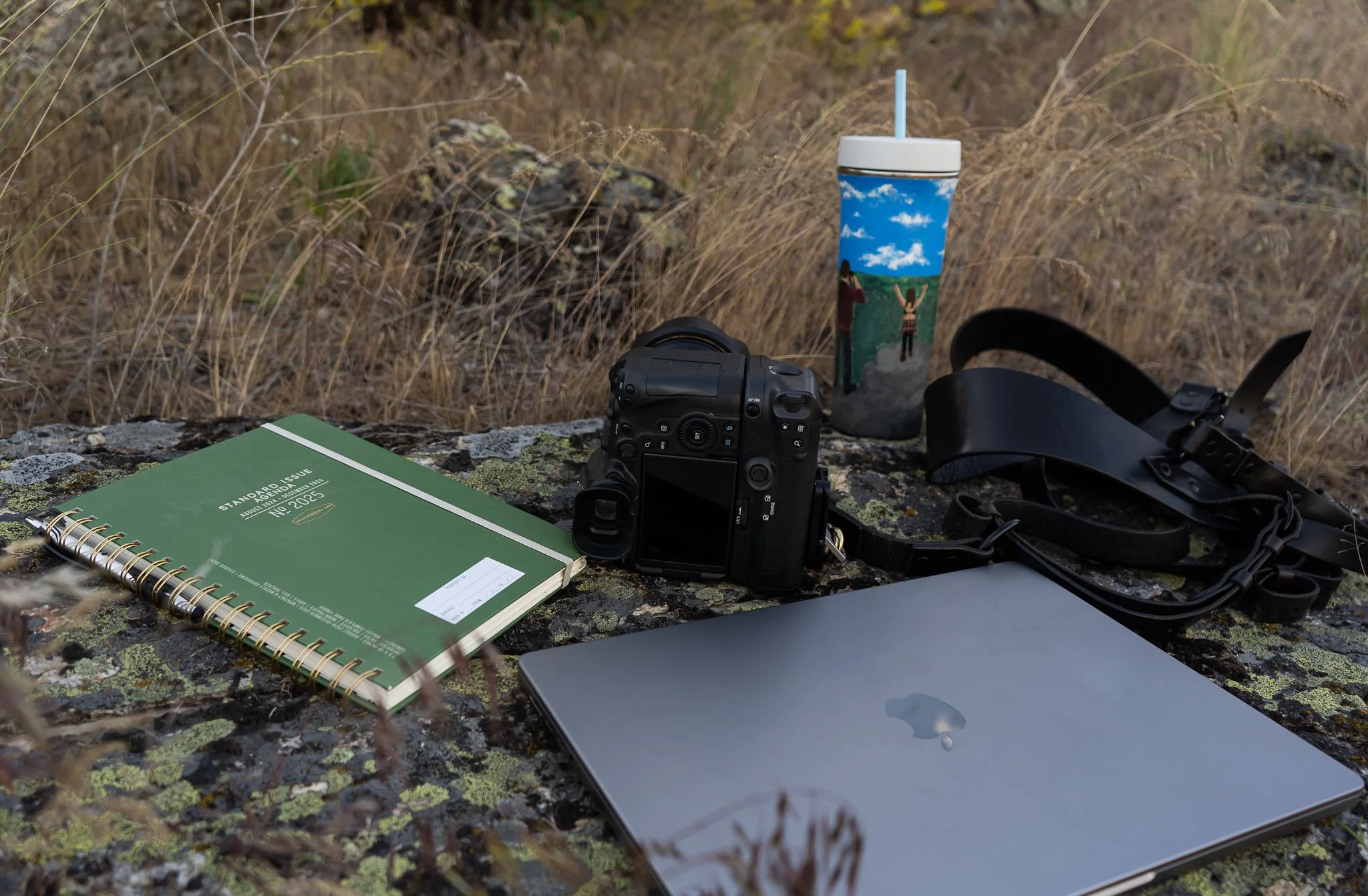 Picture of a camera, laptop, calendar book, and tumbler all sitting on a big rock