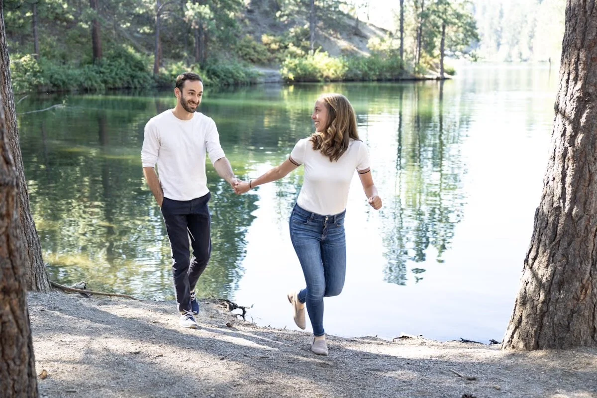A man and woman holding hands and smiling at each other near a lake in a forest.