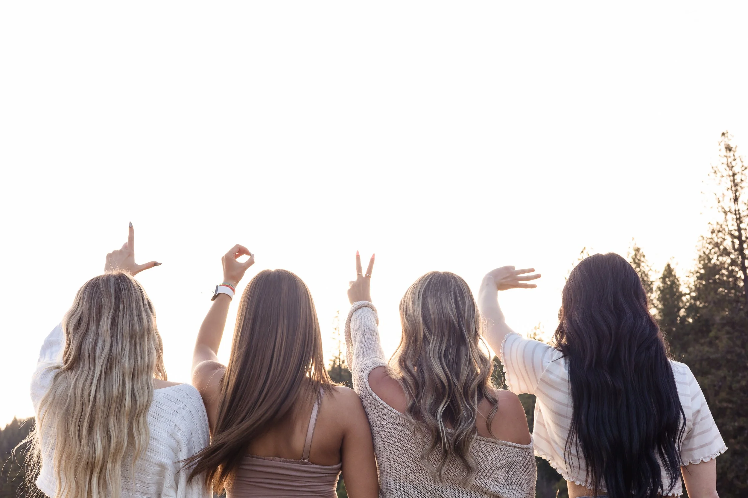 Five young women with long hair stand outdoors during sunset, with trees in the background, and are making hand gestures.