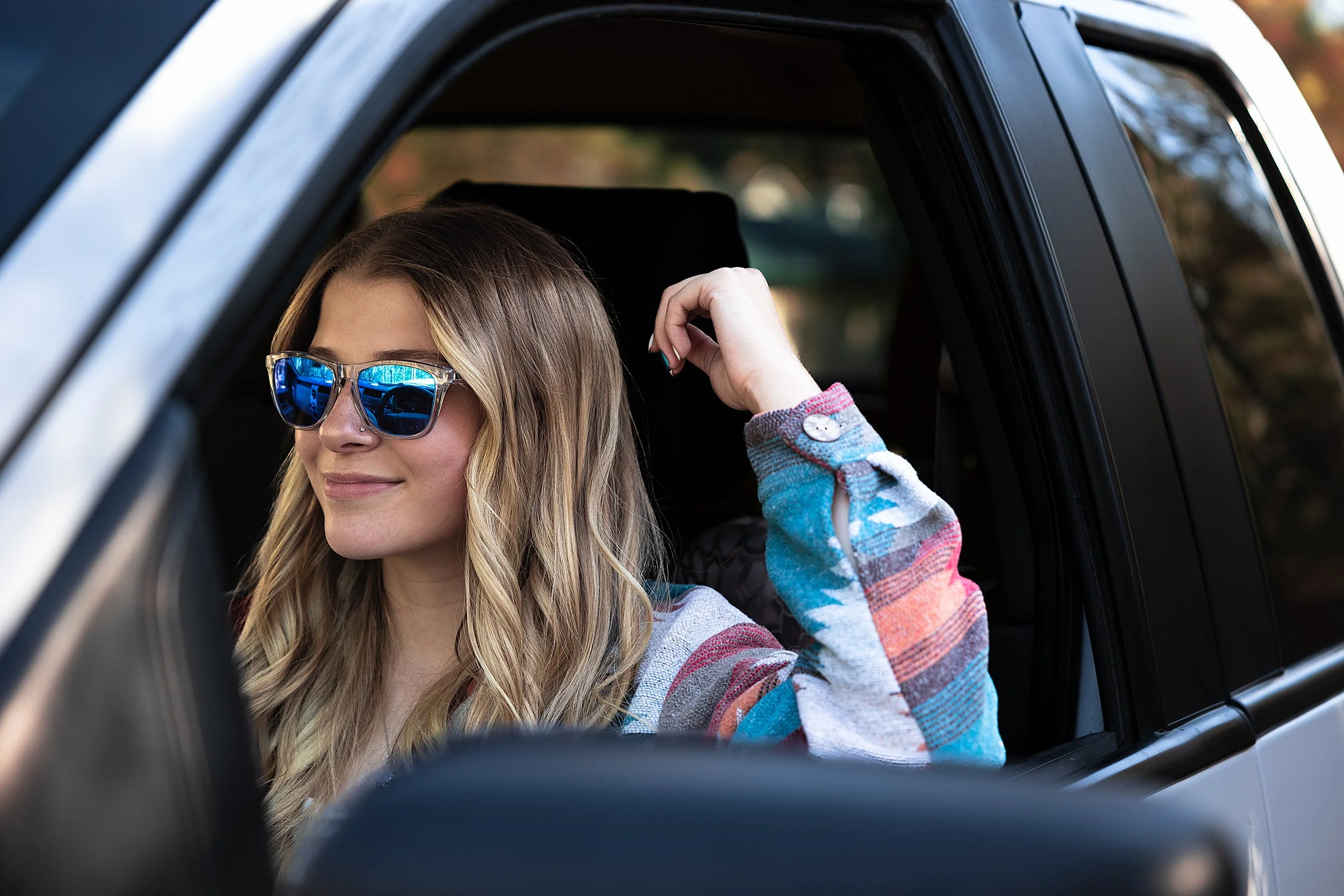 A woman with wavy blonde hair wearing blue reflective sunglasses and a colorful plaid shirt sitting inside a car.