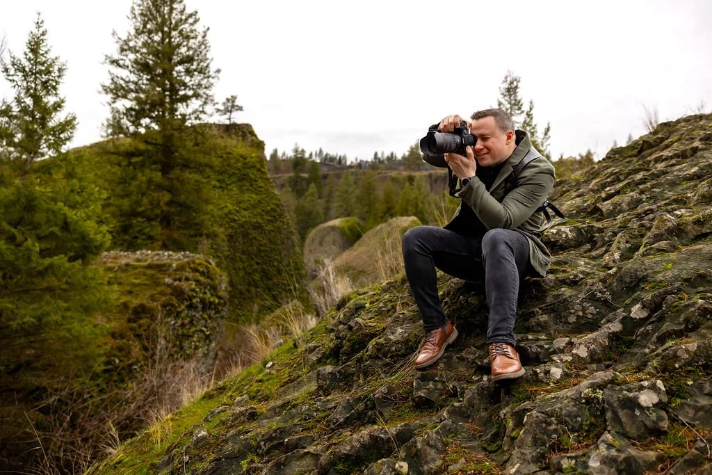 A man sitting on a rocky ledge during a hike, holding a camera and smiling while enjoying the view of trees and the distant landscape.
