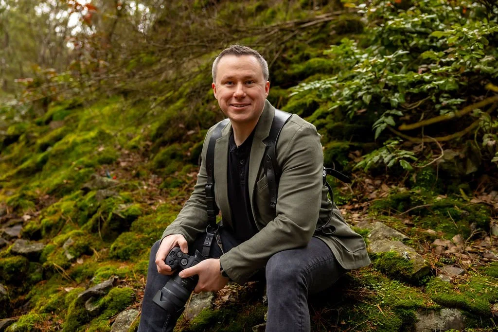 Man sitting on mossy rocks in a forest holding a camera, smiling at the camera.