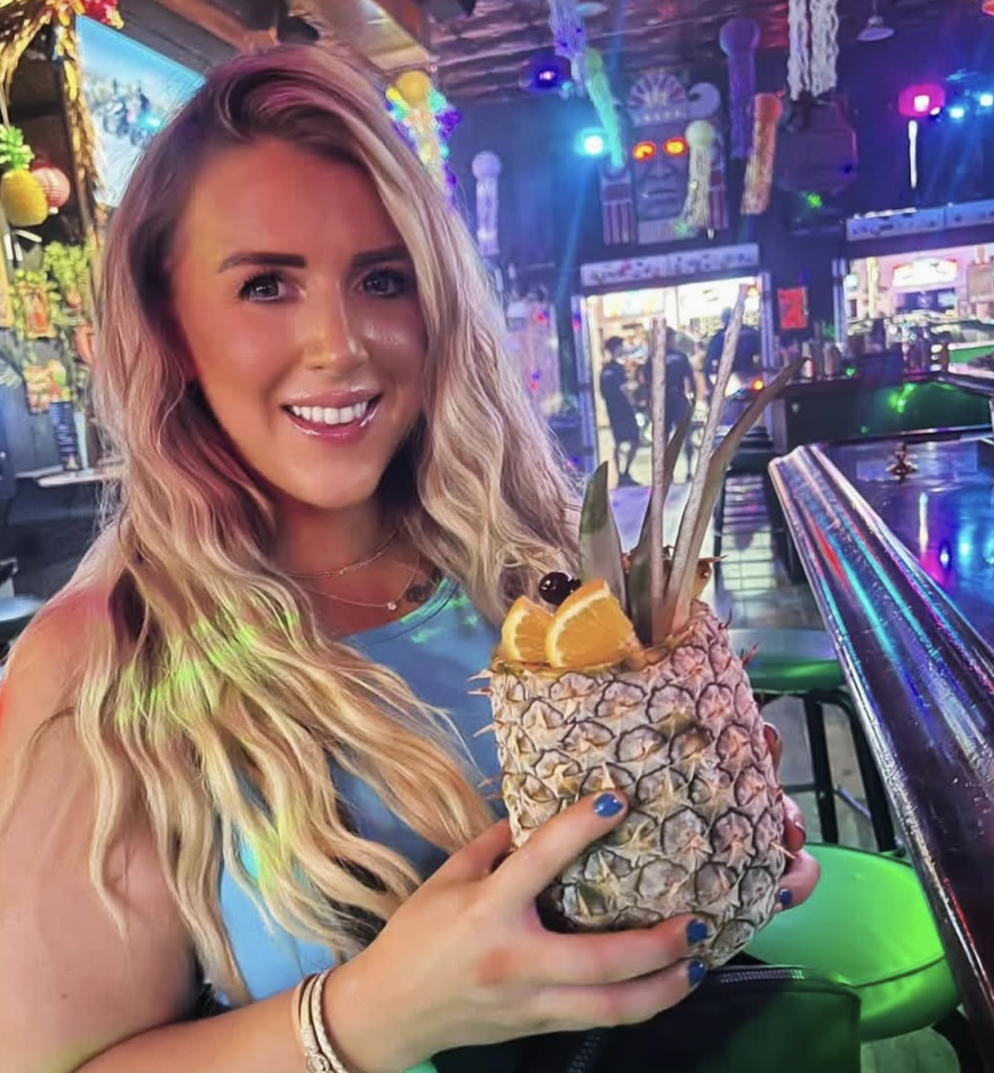A smiling woman with long, wavy blonde hair holding a pineapple-shaped cocktail with fruit garnishes in a colorful, lively bar or club setting with neon lights and decorations.