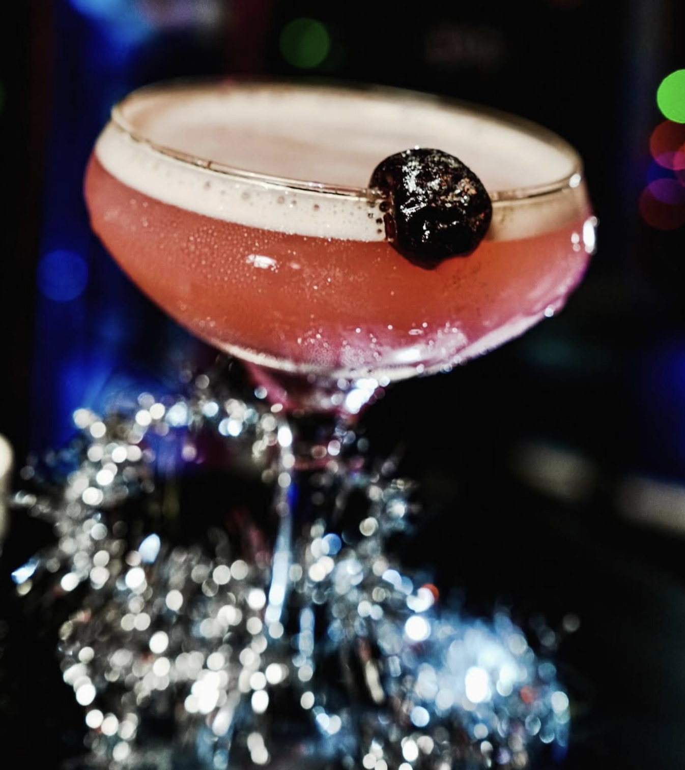 A pink cocktail in a coupe glass garnished with a dark cherry, with colorful blurred lights in the background.