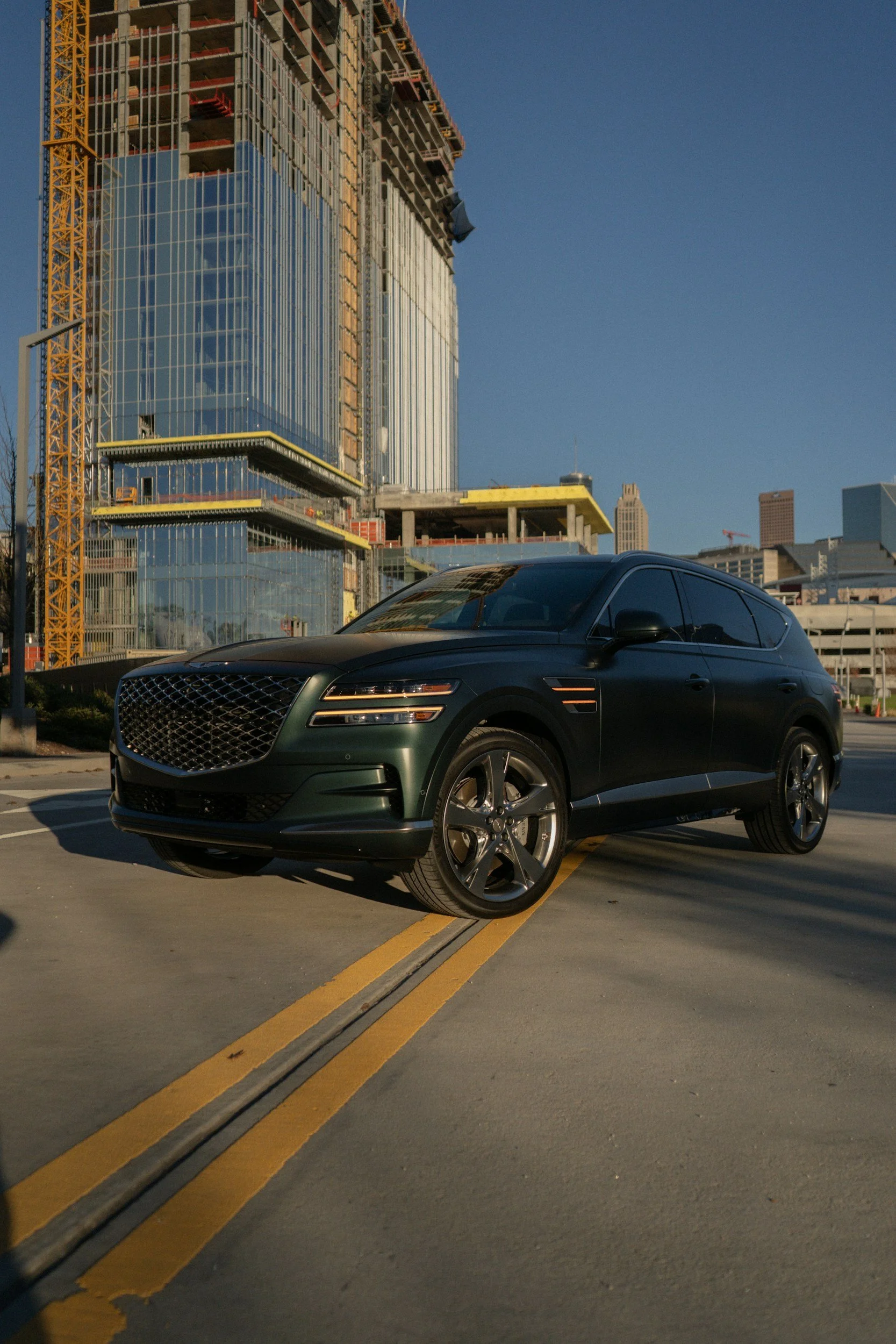 A sleek, dark green SUV is parked on a city street, with a construction site and modern glass buildings in the background during daylight.