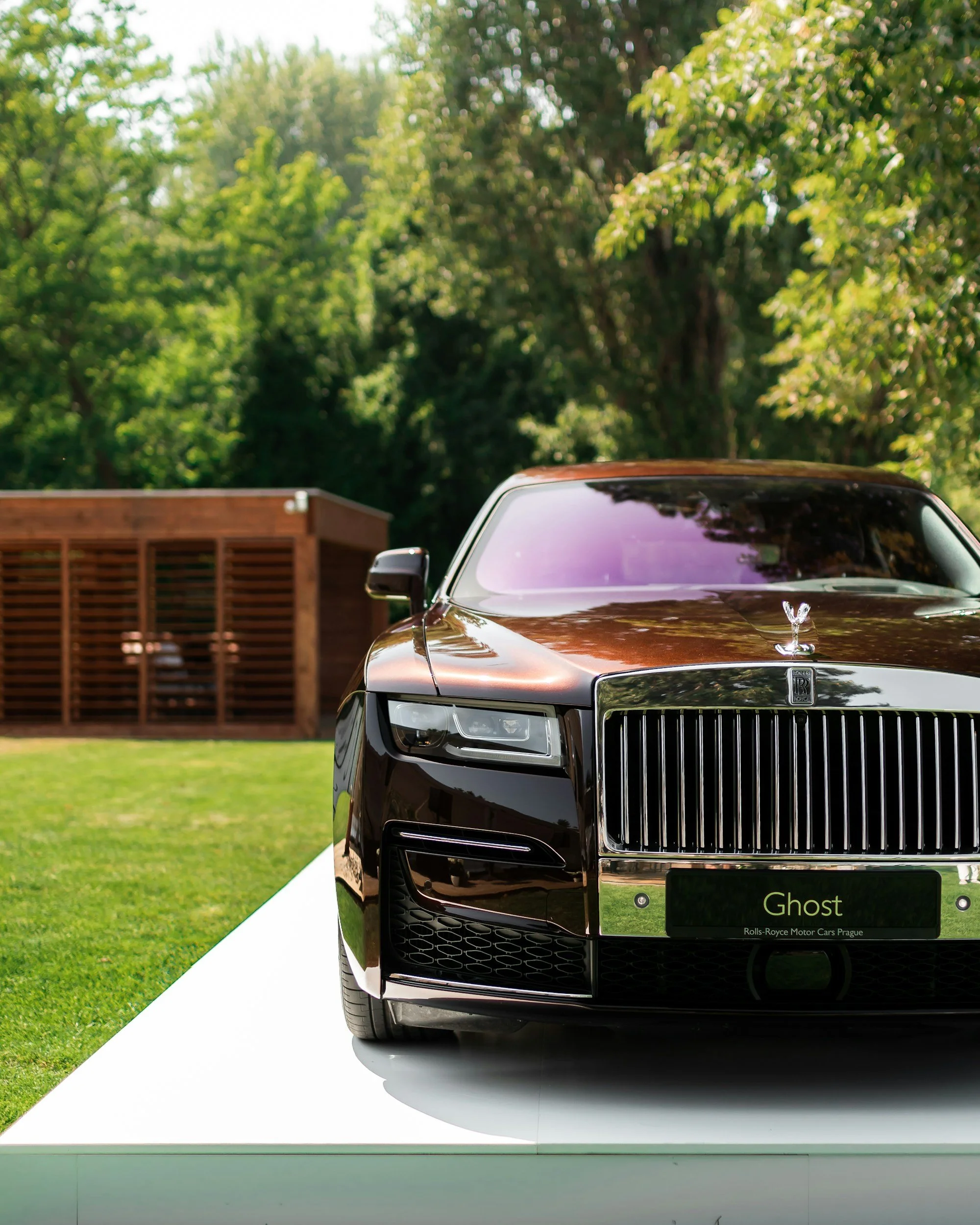 Close-up of a black Rolls-Royce Ghost car displayed outdoors on a white platform, with trees and a wooden structure in the background.