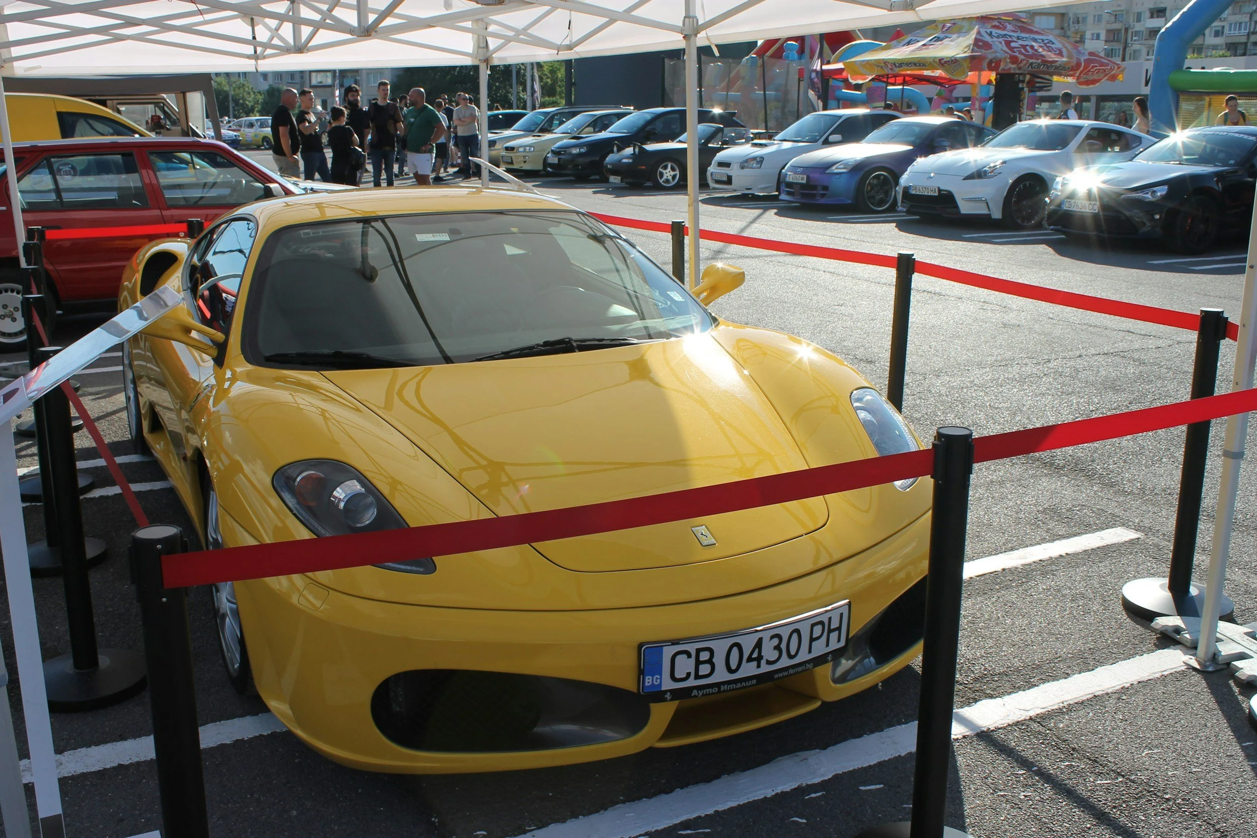 A yellow Ferrari sports car on display at an outdoor car show, surrounded by a red rope barrier. There are other cars and a crowd of people in the background under a white tent canopy.