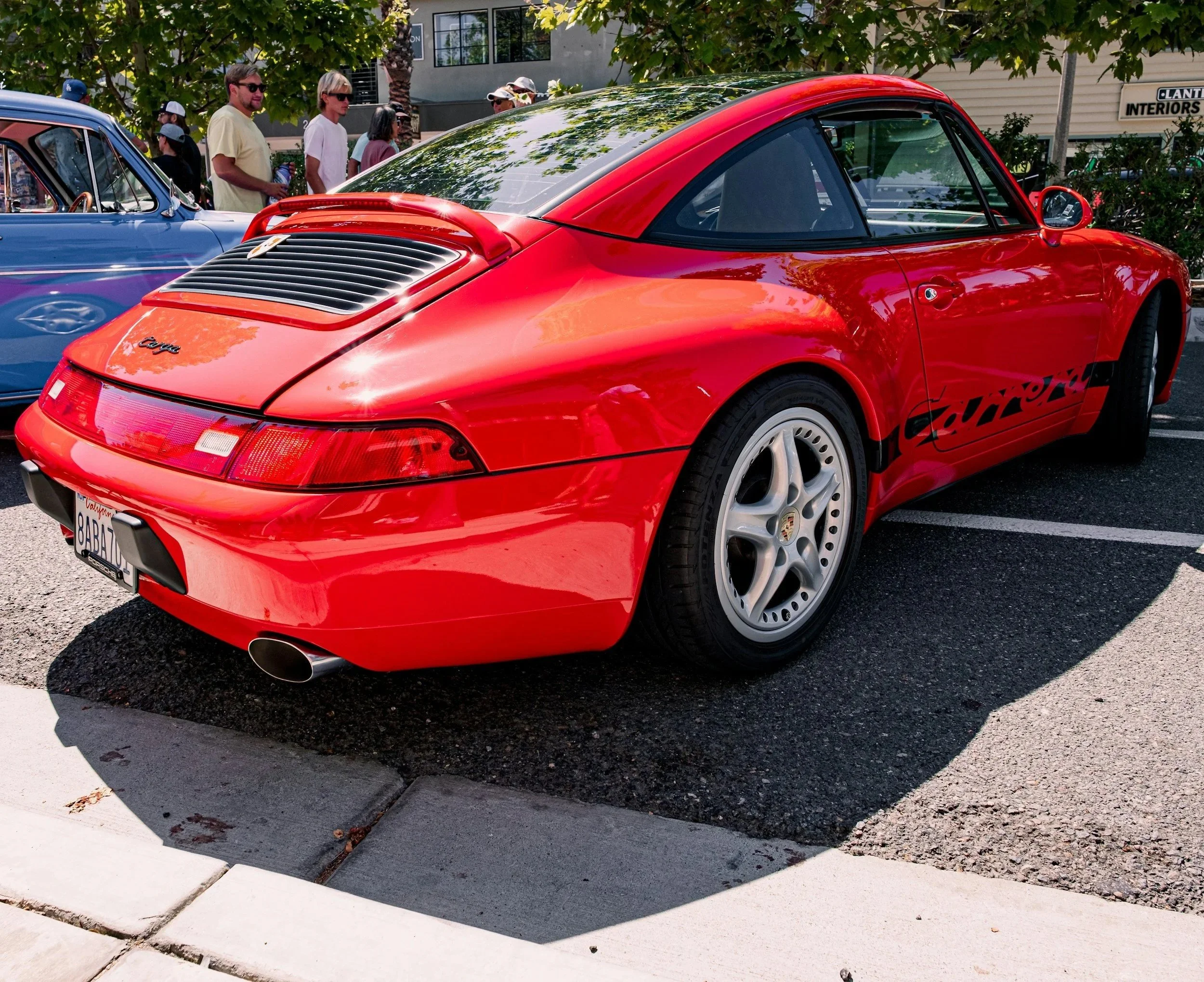 Red Porsche Carrera parked in a parking lot during a car show, with people and trees in the background.