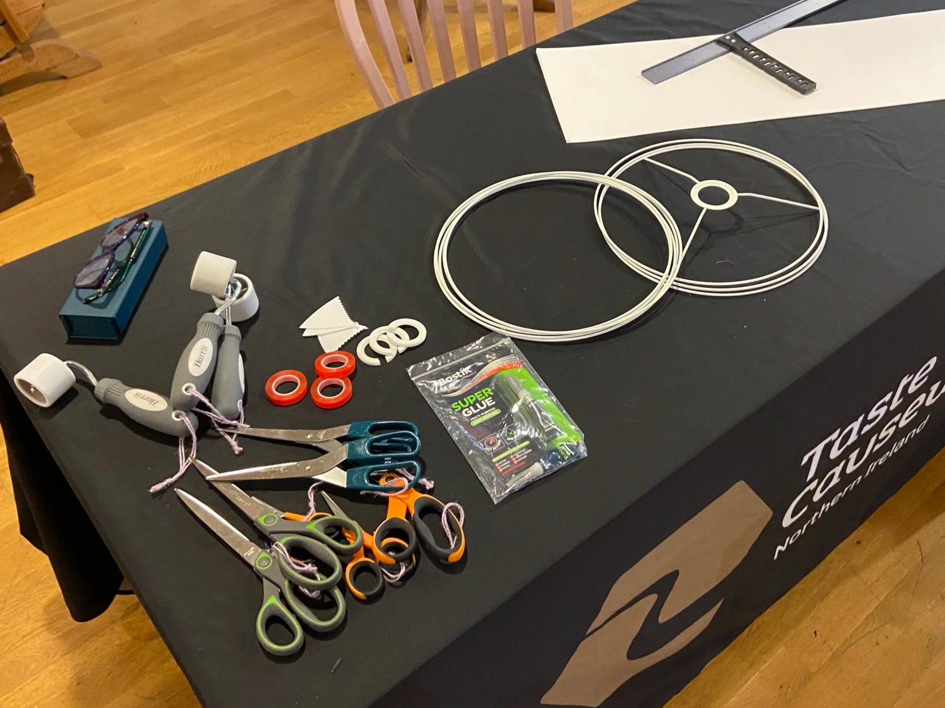 A table covered with lampshade making crafting supplies, including scissors, and a pack of super glue, with a pair of glasses, a box, and some rulers.