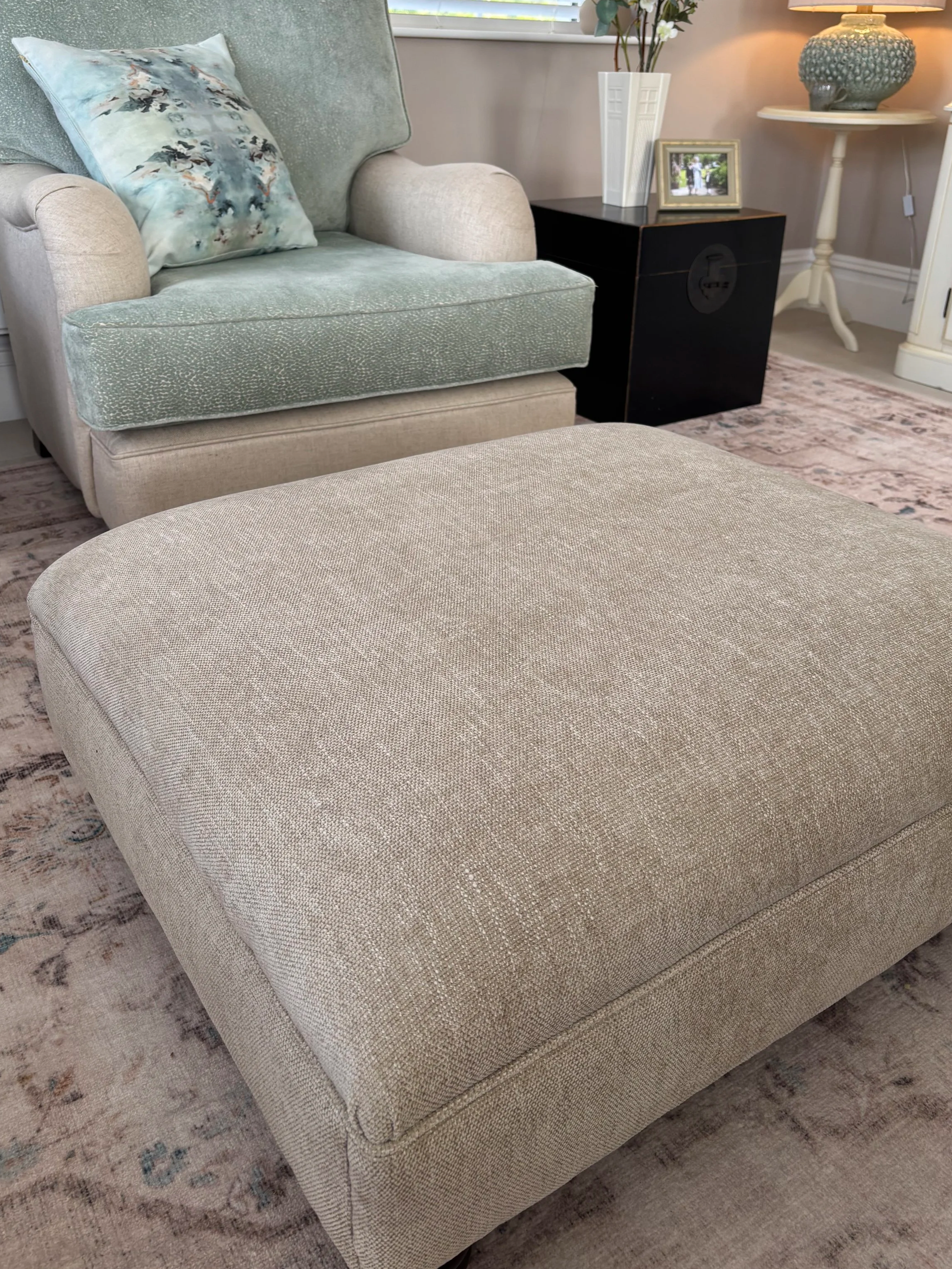 Living room with beige textured ottoman in the foreground, light green armchair with a matching textured cushion, a side table with a white vase with white flowers, a picture frame, and a decorative lamp on top. A window with blinds and a patterned rug are also visible.
