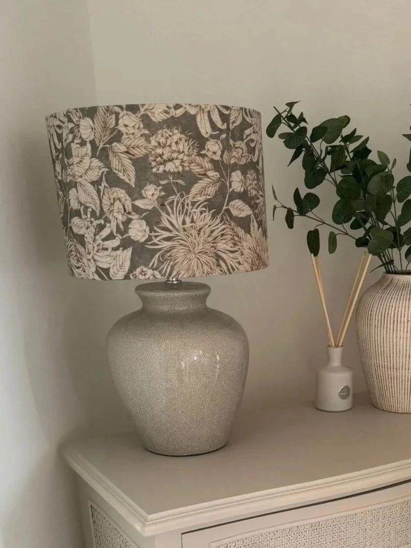 A decorative table lamp with a floral patterned lampshade, placed on a white side table next to a potted green plant and a small white vase with reed diffusers. neutral in colour. made by a student during a Purple Heather Lampshade workshop
