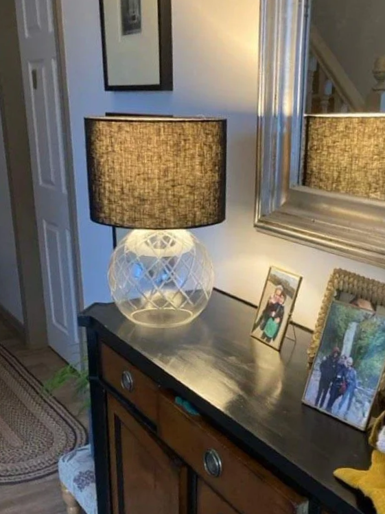 A sideboard with framed photos, a large mirror, and a table lamp with a textured glass base and a dark fabric shade. lampshade handmade by a person who attended a lampshade making workshop 