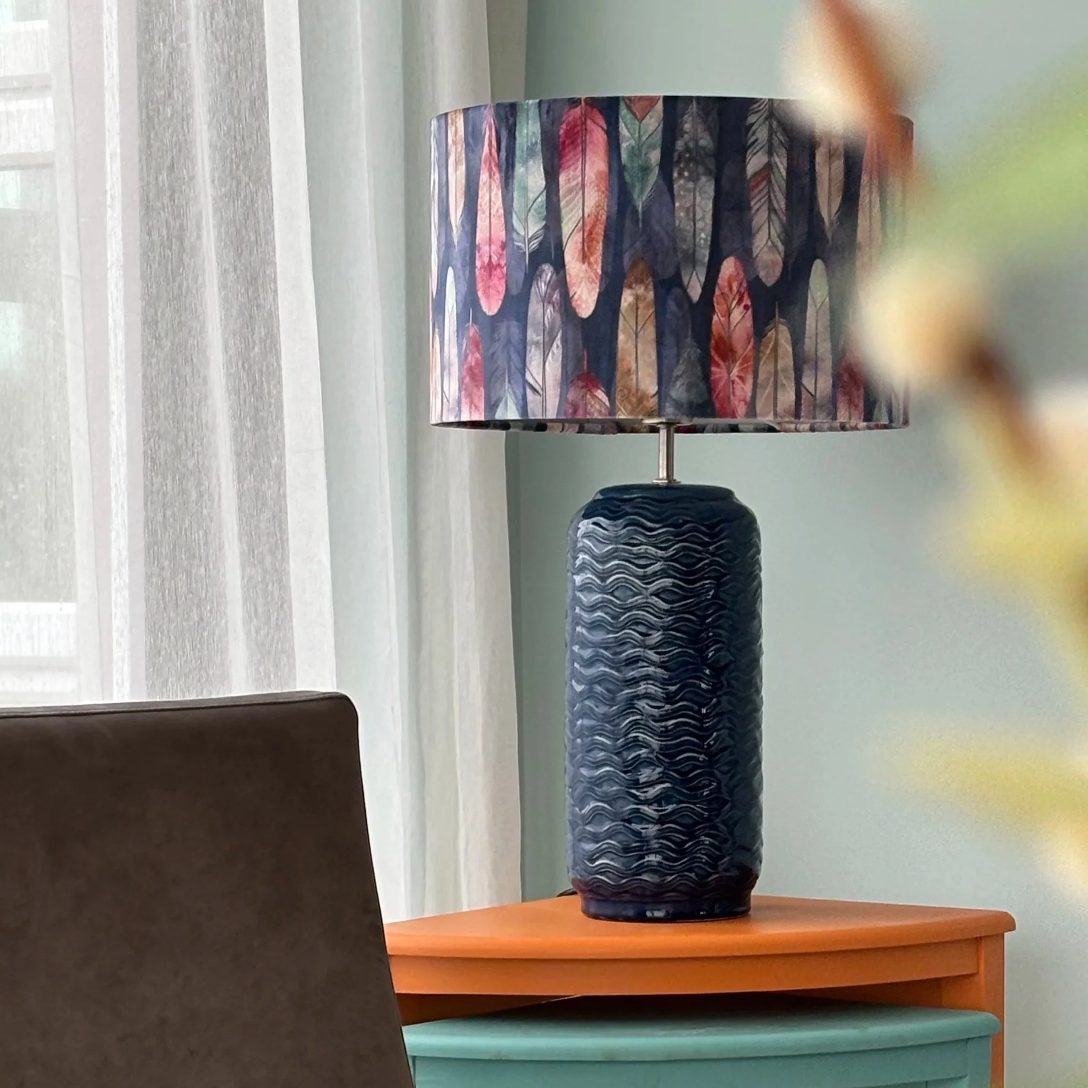 A colourful table lamp with a feather pattern on the lampshade, placed on a wooden table. colourful bespoke velvet drum shaped lampshade set on a ceramic blue table lamp base