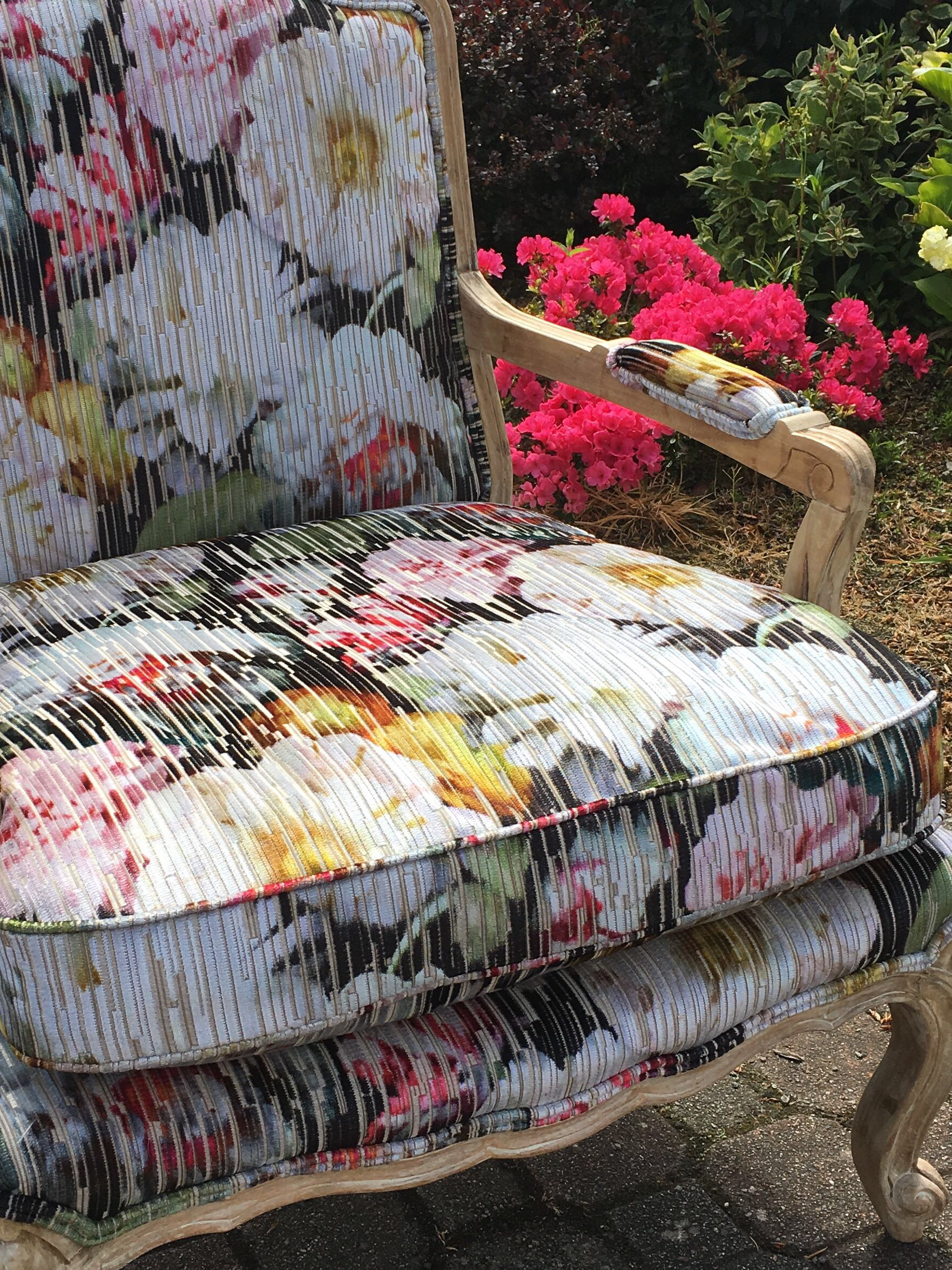 A vintage wooden outdoor bench with floral-patterned fabric upholstery on the seat and backrest, surrounded by lush pink and white flowers and green foliage.
