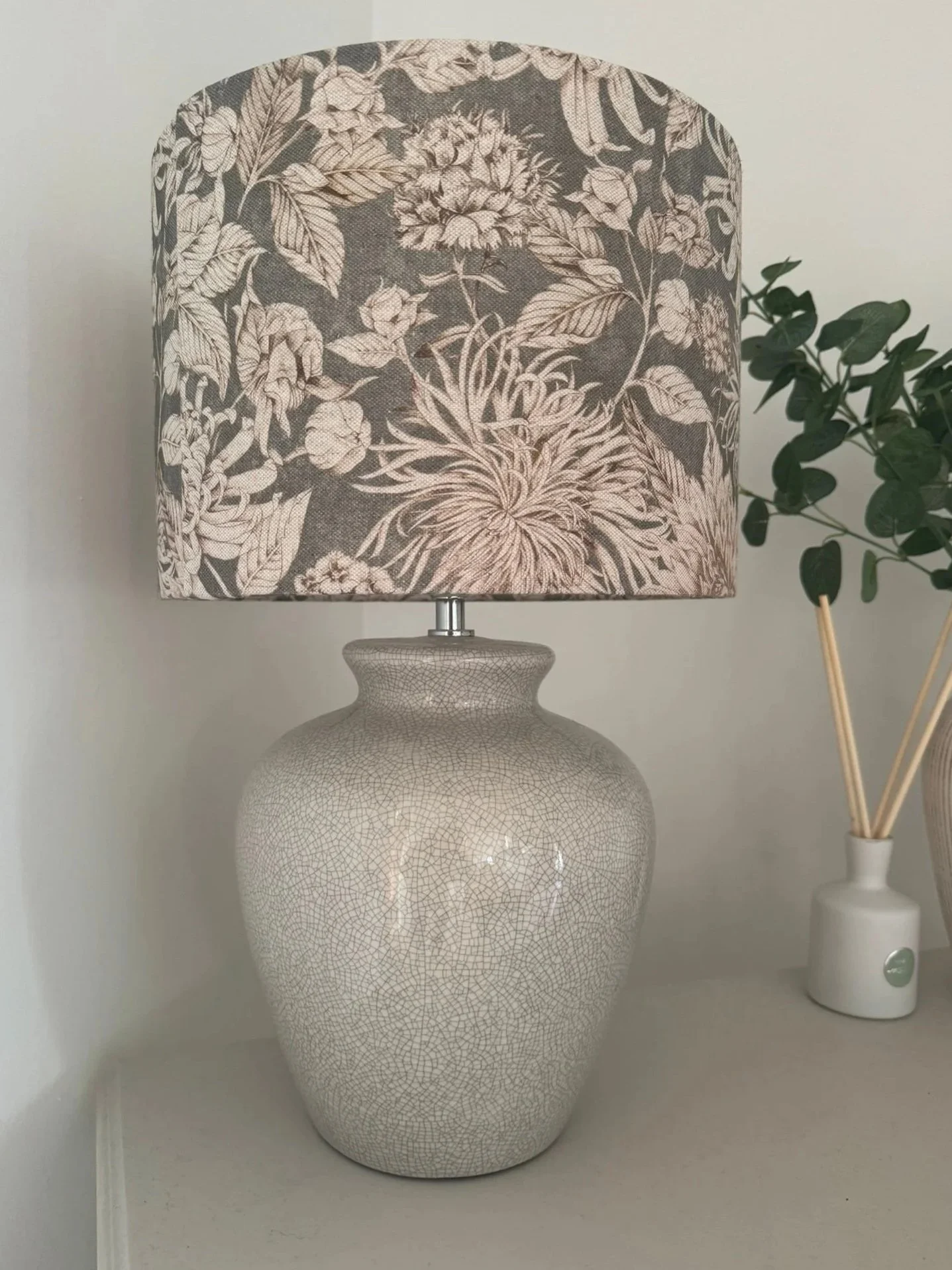 A ceramic table lamp with a crackled finish and a floral patterned fabric lampshade, placed on a white surface. Next to it, a white vase holds wooden sticks and a green leafy plant. lampshade handmade by a person who attended a lampshade making works