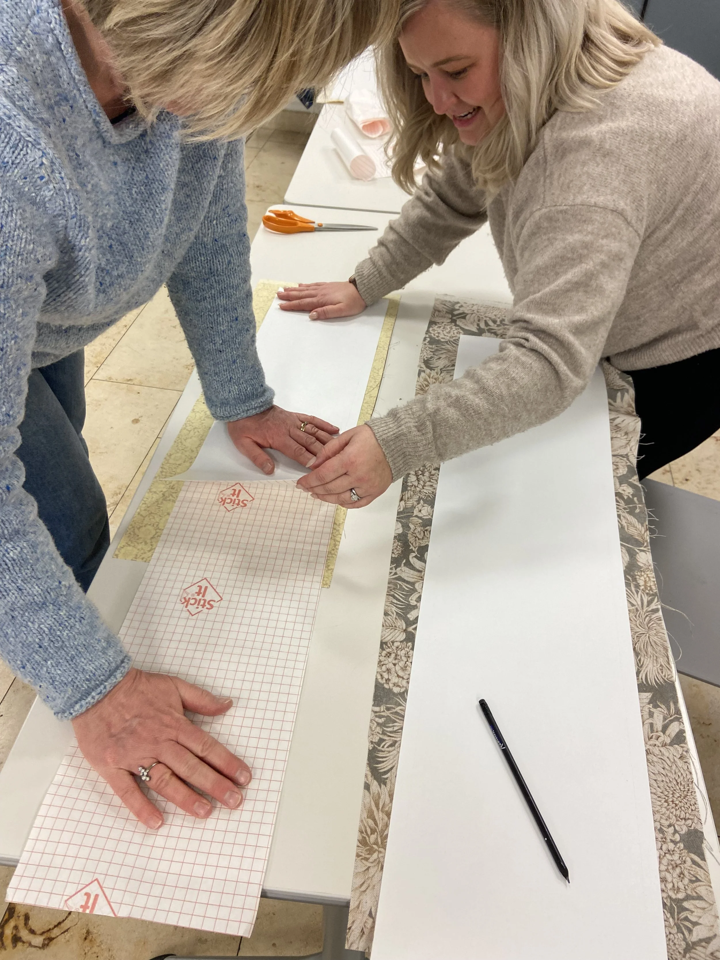 Two women are working together to make a lampshade at a table, measuring and cutting fabric with scissors, with fabric, rulers, and pens visible on the table.