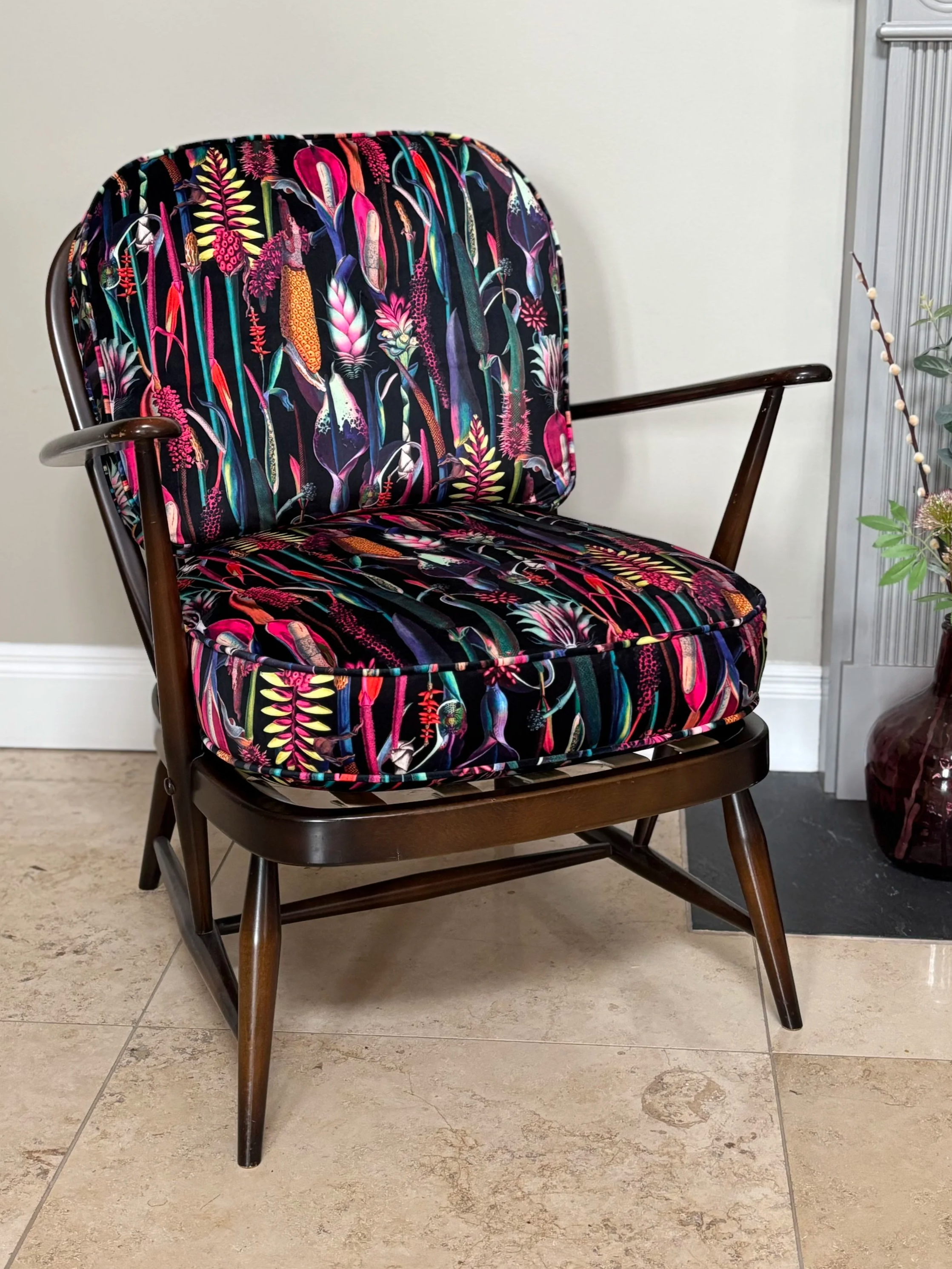 A wooden ercol vintage chair with dark arms and legs, and a cushion featuring a colourful botanical pattern with pink, yellow, and blue plants on a black background, the fabric is velvet. placed on a tiled floor against a cream-coloured wall.