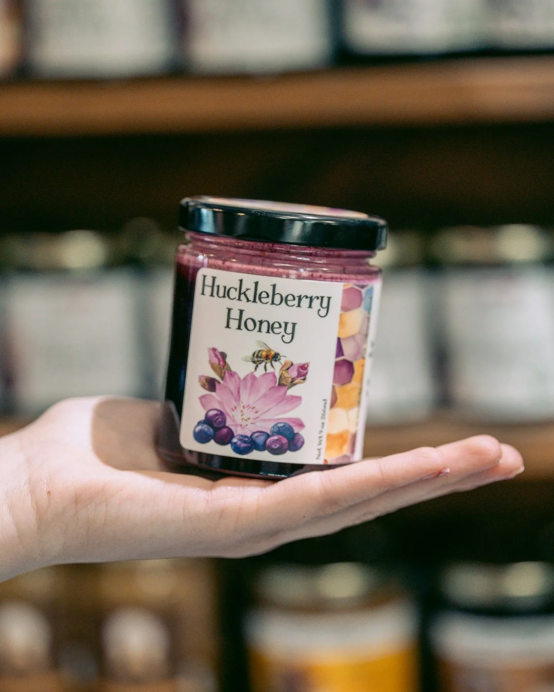 A jar of Key to the Mountains' Huckleberry Honey sits in the palm of someone's hand, with shelves of product in the background.
