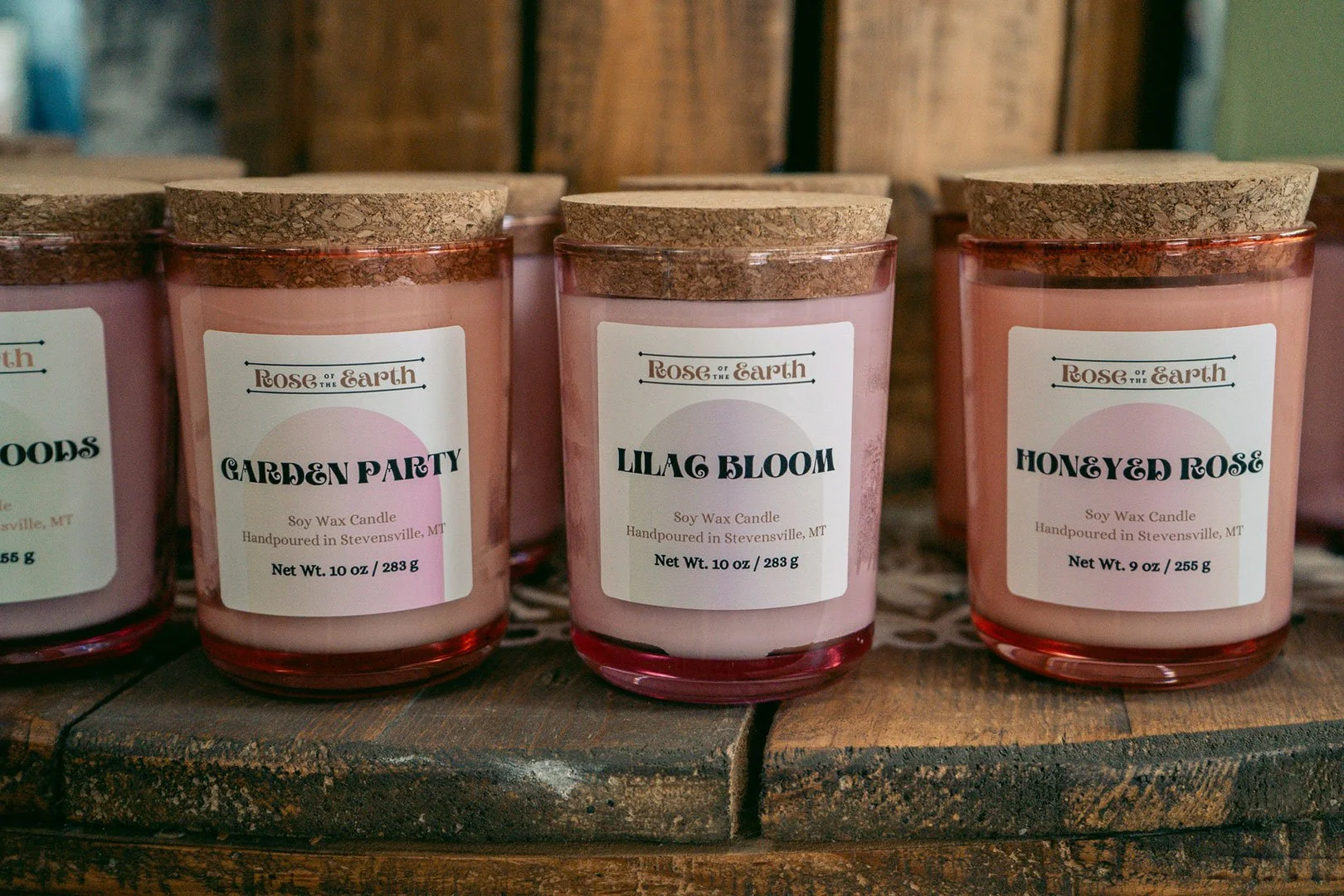 A variety of candles in brightly colored pink jars with cork toppers sits upon a rustic tabletop. Scents include Lilac Bloom, Honeyed Rose, and Garden Party.