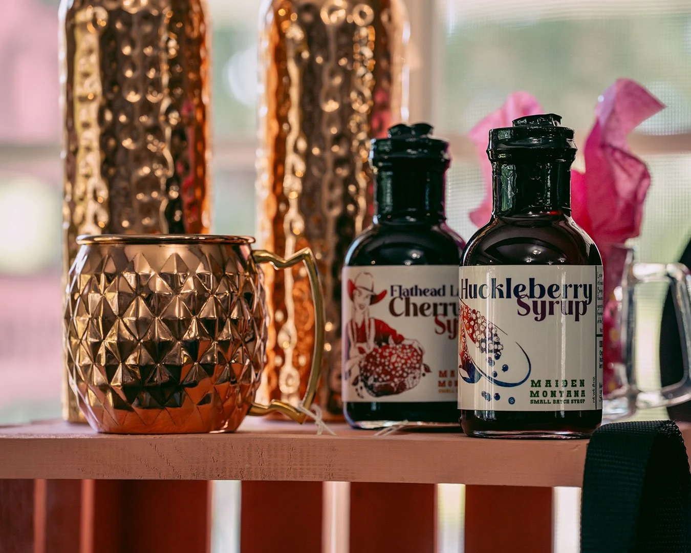 Bottles of Huckleberry & Cherry Syrups from Key to the Mountain sit atop a pink crate with a copper mug.