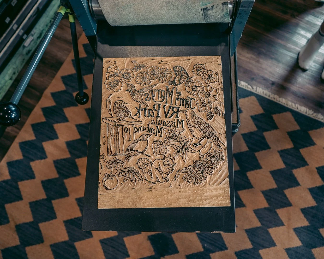 A block of wood, carved with layers of flowers and animals found  at Jim & Mary's RV Park, sits upon a large press, ready for printing..