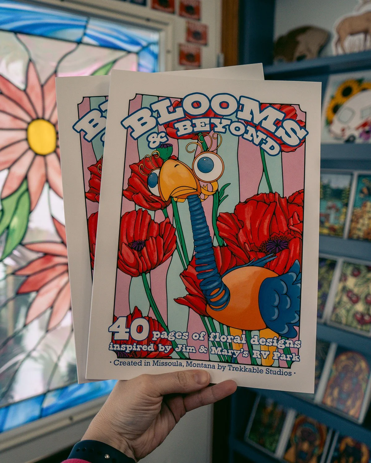 Two Blooms & Beyond Coloring Books are held up, showing off the brightly colored cover featuring a decorative bird statue and bright red poppies.