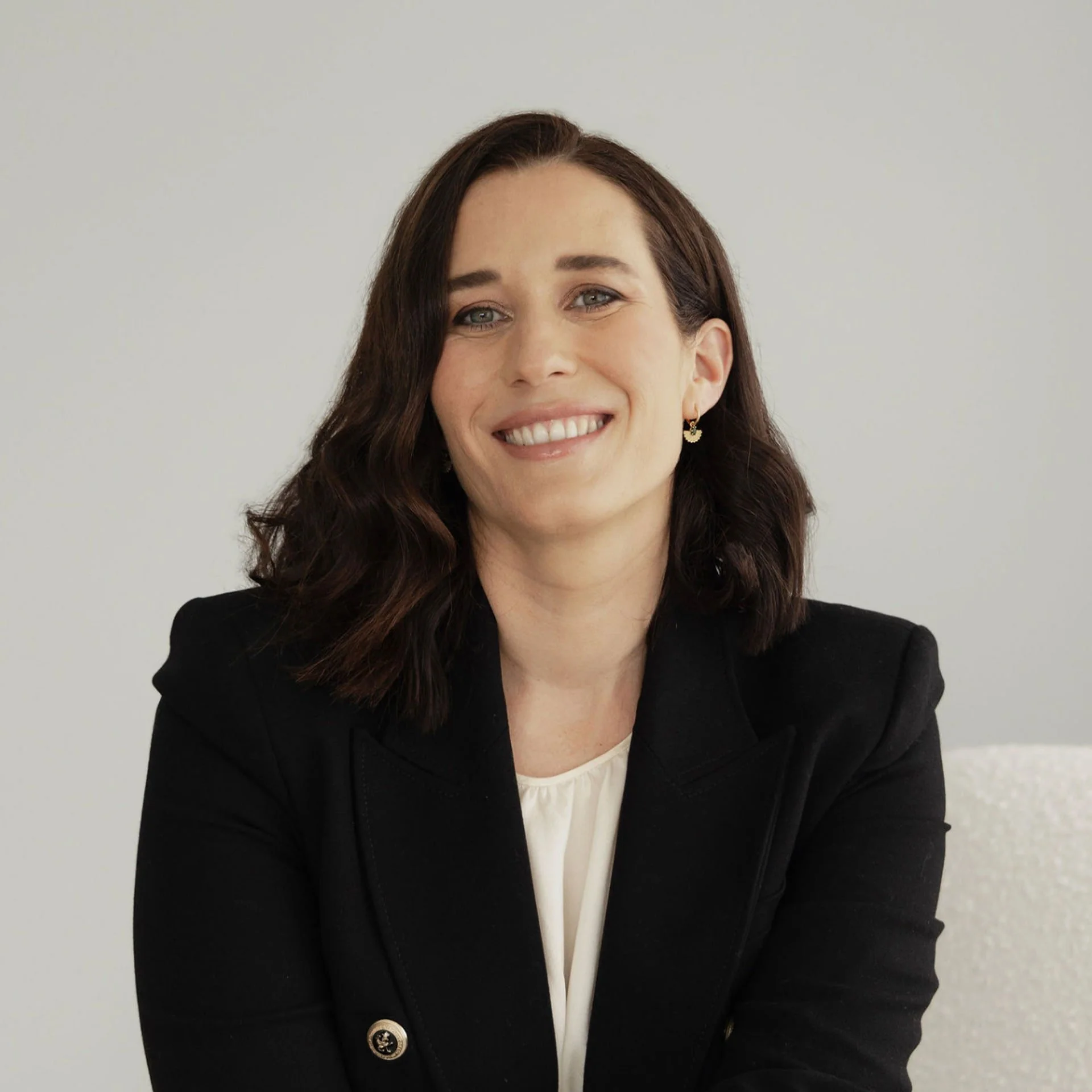 Close-up of a smiling woman with shoulder-length brown hair, wearing a black blazer and a white top, sitting against a plain light background.