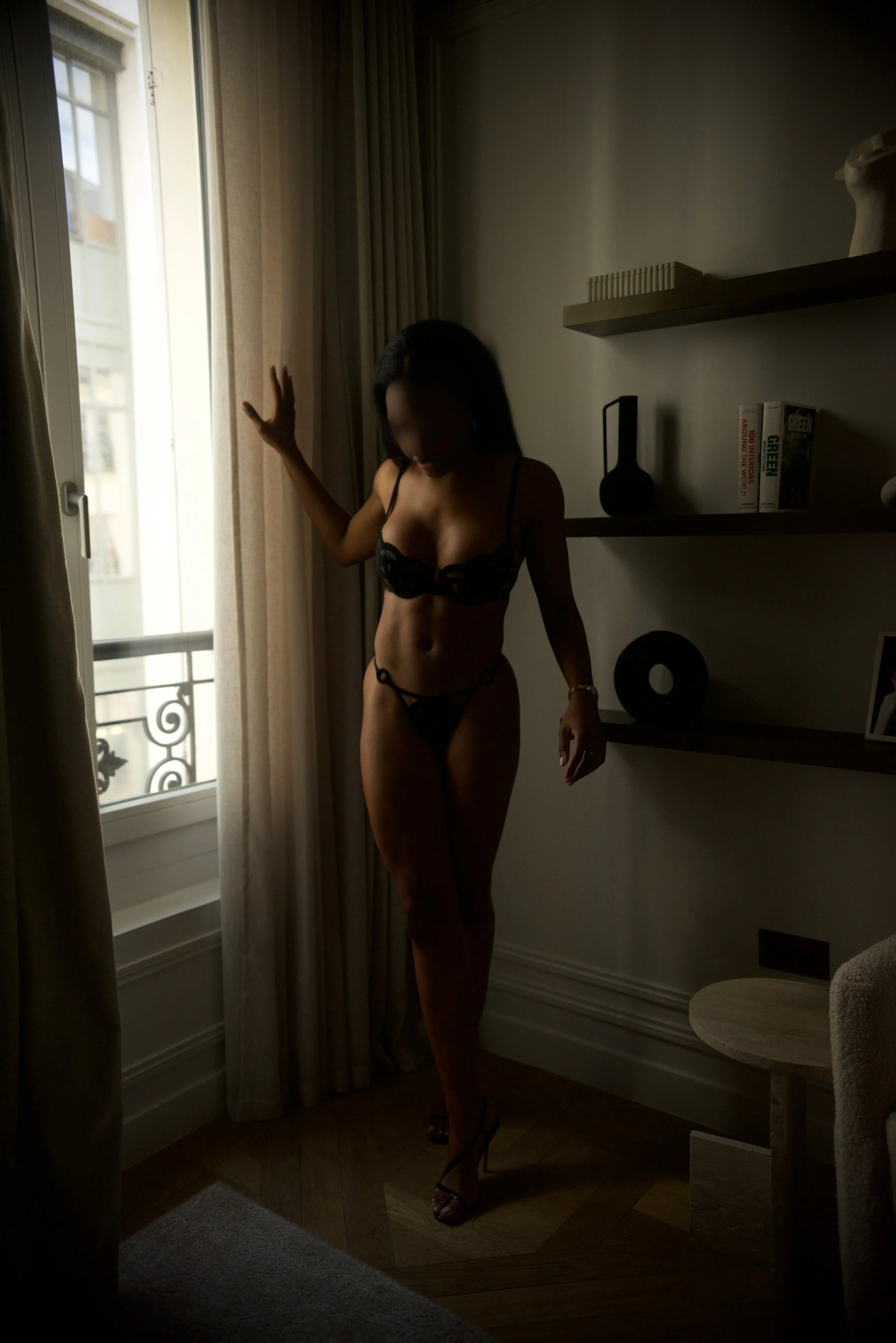 Femme en sous-vêtements noirs, debout près d'une fenêtre avec des rideaux, dans une pièce sombre avec une étagère contenant des livres et des décorations.