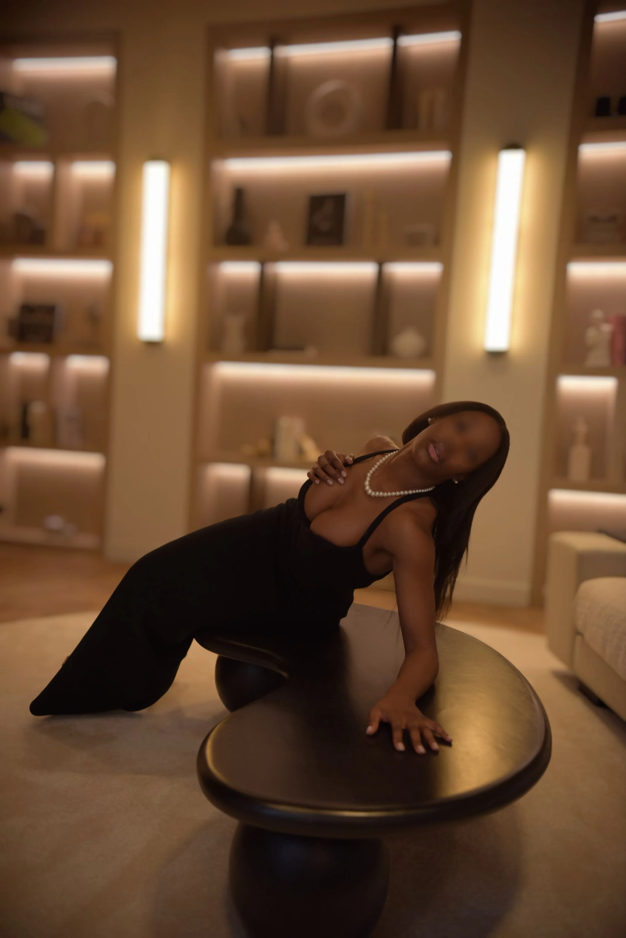 Une femme élégante portant une robe noire et un collier de perles pose sur une table de salon dans une pièce lumineuse avec des étagères en arrière-plan.