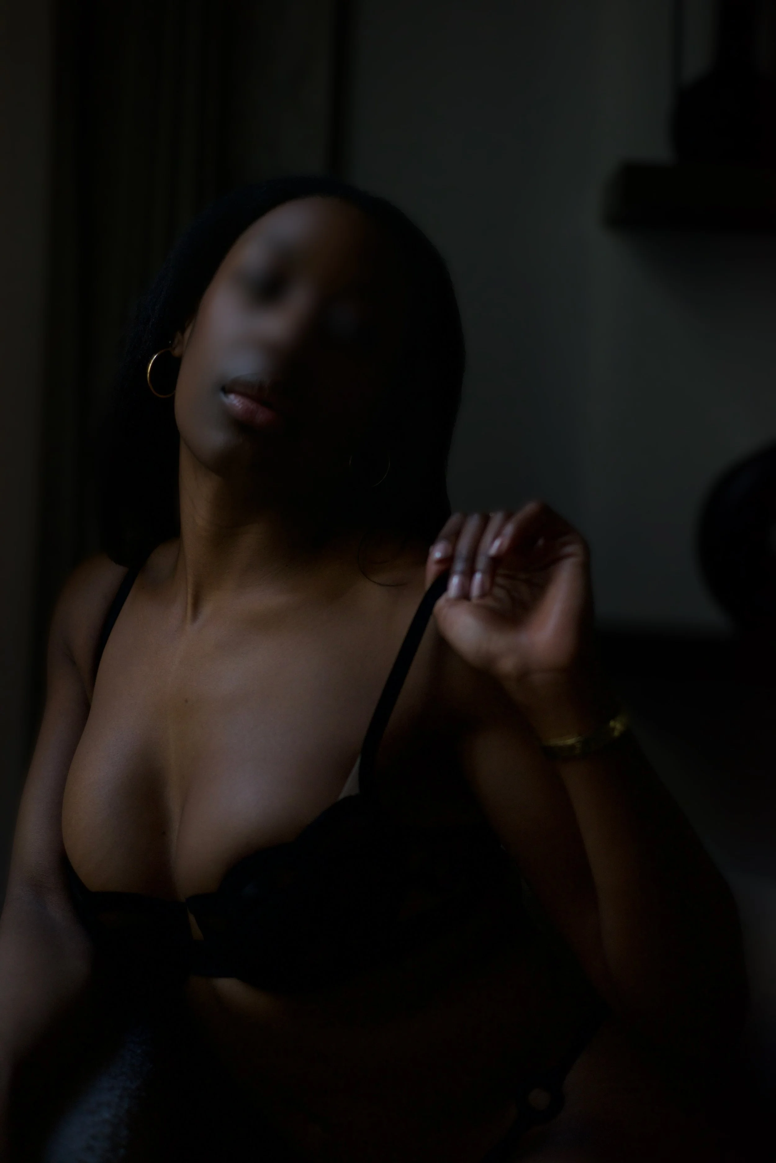 Une femme portant un haut noir à épaules dénudées, avec des boucles d'oreilles en or, dans une pièce sombre, posant près d'une fenêtre.